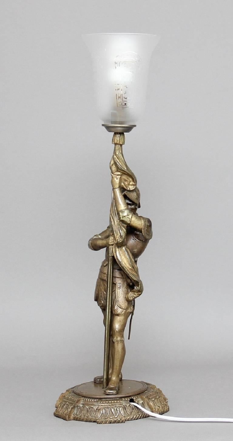 Early 20th century brass lamp of a figure of a knight holding a flag pole standing on a circular base, with the top of the flag pole supporting a glass shade, circa 1920.