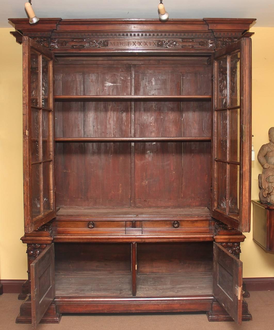 A large highly carved 19th century Italian walnut bookcase or cabinet, the moulded cornice above a carved frieze, with two large glazed doors opening to reveal two shelves inside, the doors flanked by carved scrolls, the lower section having four