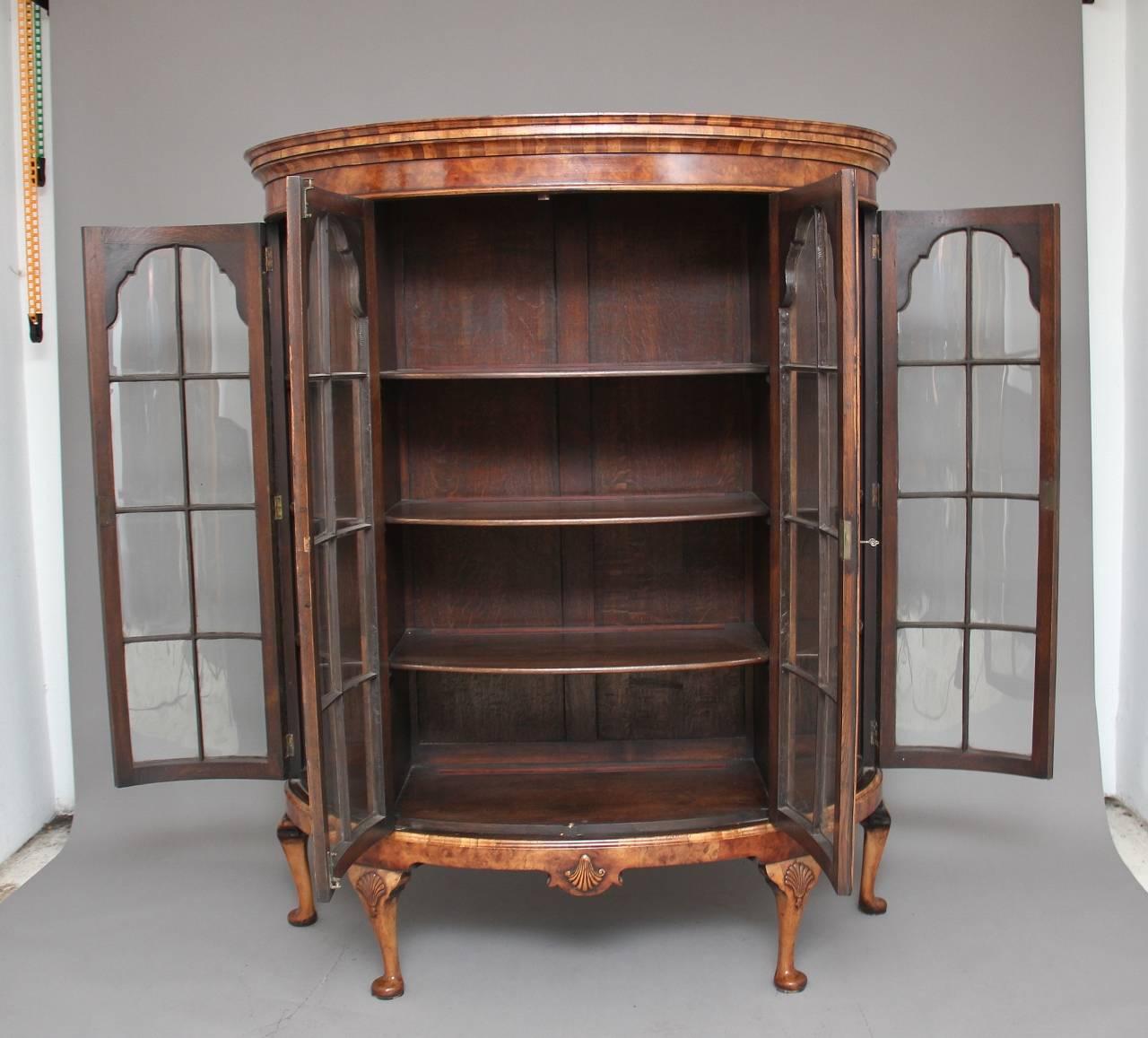 Early 20th century walnut bowfronted display cabinet, the moulded cornice above four arched glazed doors in the Queen Anne style with cross grain mouldings, each door opening to reveal three fixed oak lined shelves inside, the shaped apron below