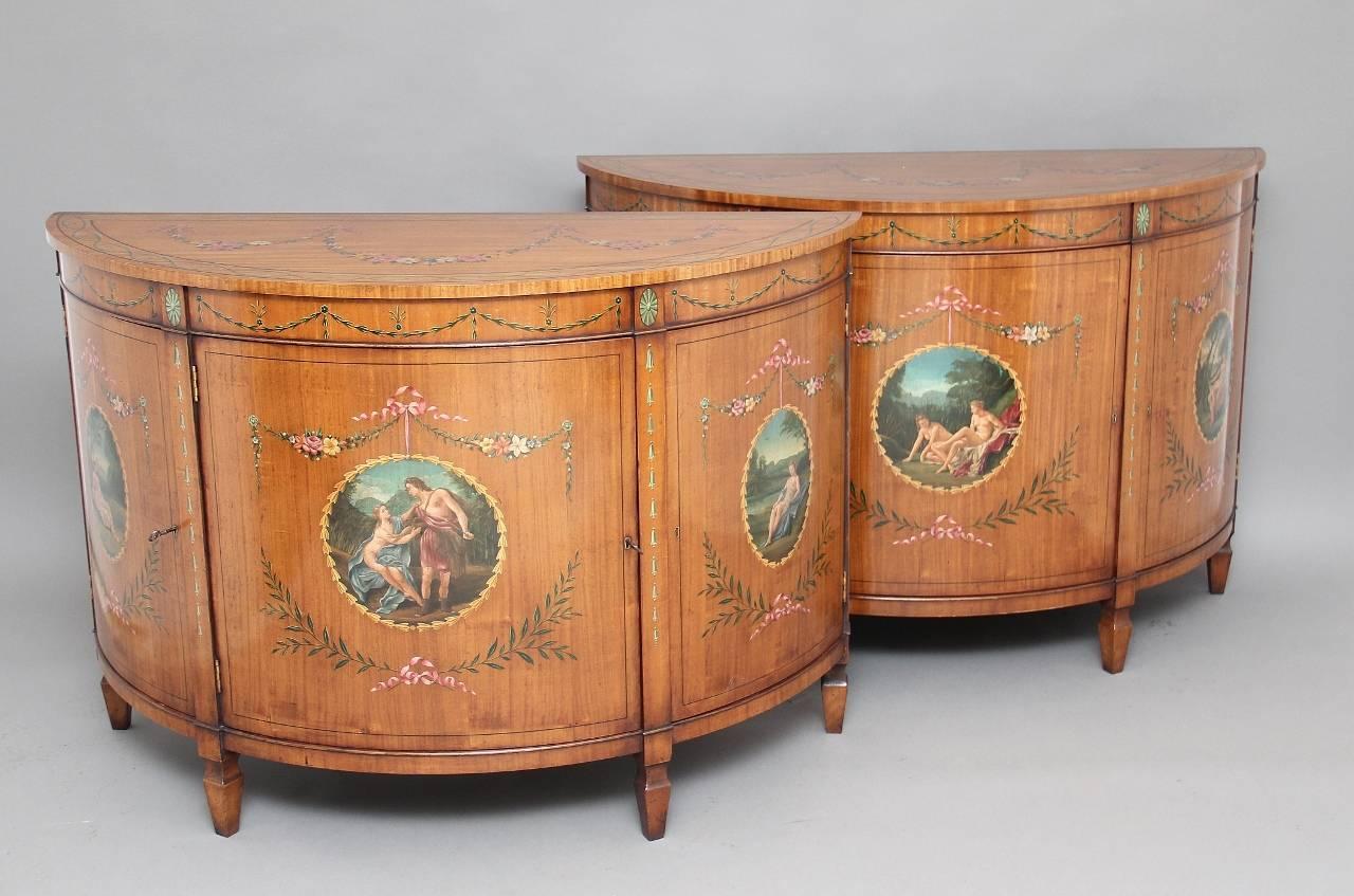 A pair of mid-20th century satinwood and painted demilune commodes in fantastic condition, the semi circular tops painted with ribbons and floral decoration with the border depicting an elegant intertwining pattern, the front of the cabinets