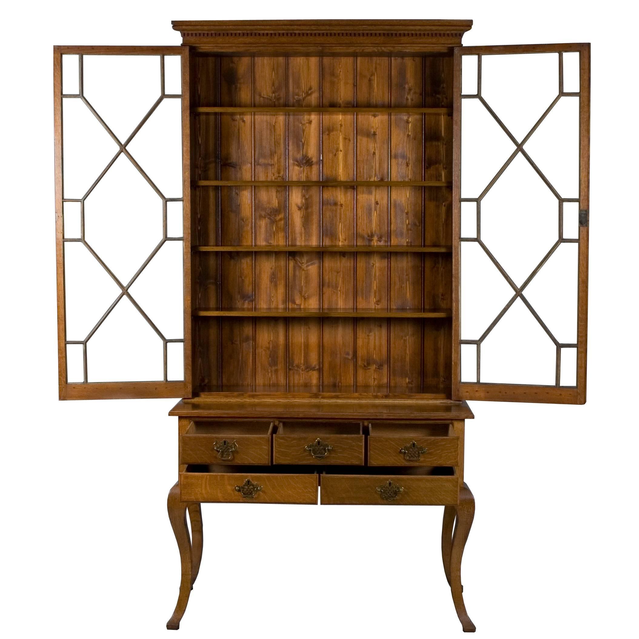 This stunning tall glass door bookcase is an English antique made circa 1890 out of oakwood. Two astragal glazed glass doors cover four adjustable wood shelves and rest upon a tall base with five drawers. The doors and drawers all lock and two keys
