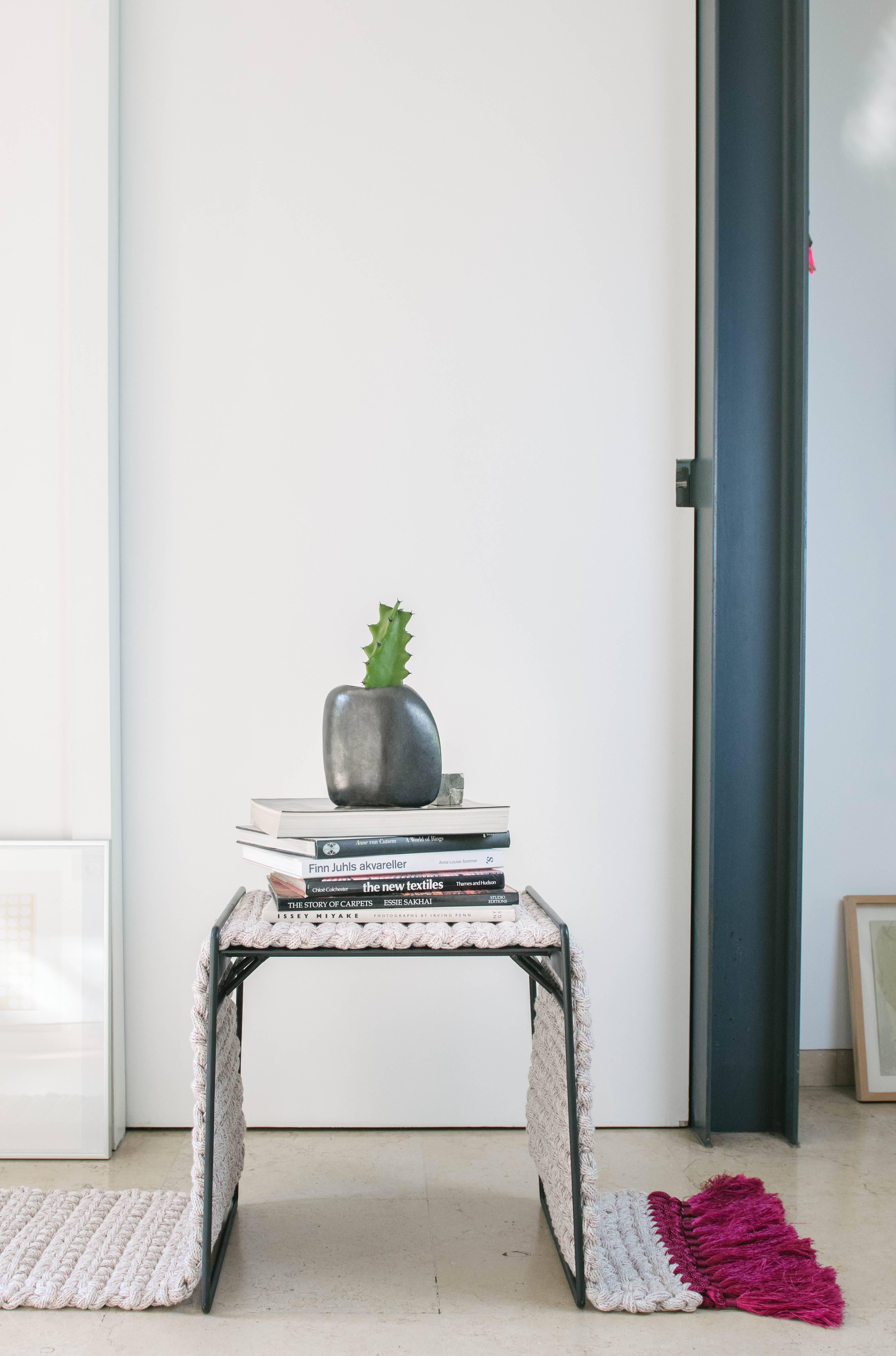 The iota rug stool is a matte black iron stool covered by a white and fuchsia tailored rug for comfortable sitting. The rug can be imaginatively arranged in different shapes which create altering appearances and different space occupations. The rug