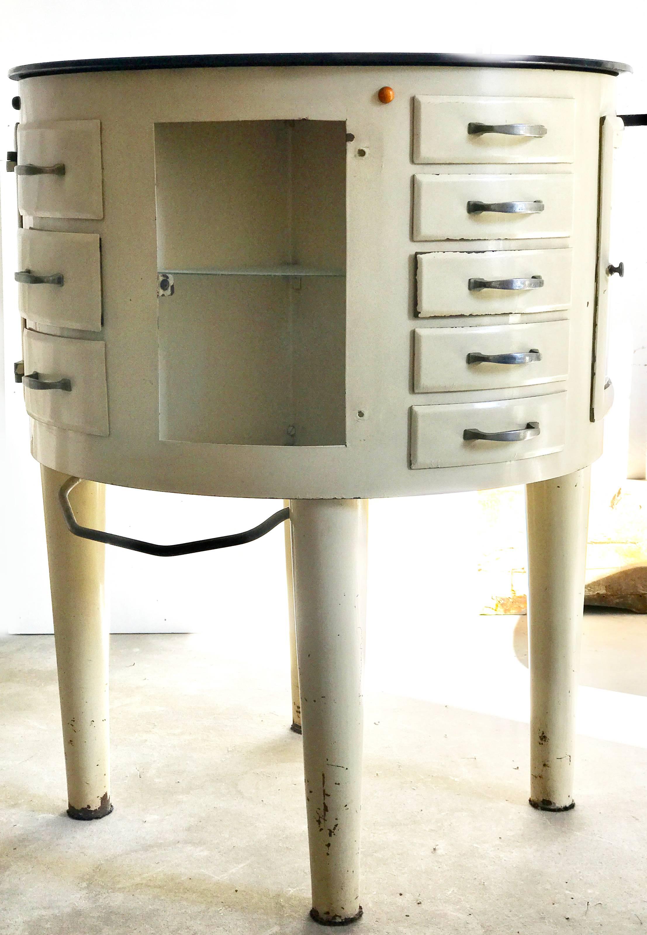 Piece of dentist furniture with a black glass revolving countertop and enameled iron storage unit that is also rotatory. It has 3 units of drawers one with 3 drawers and two with five drawers, all of them have glass interior and 3 units with one