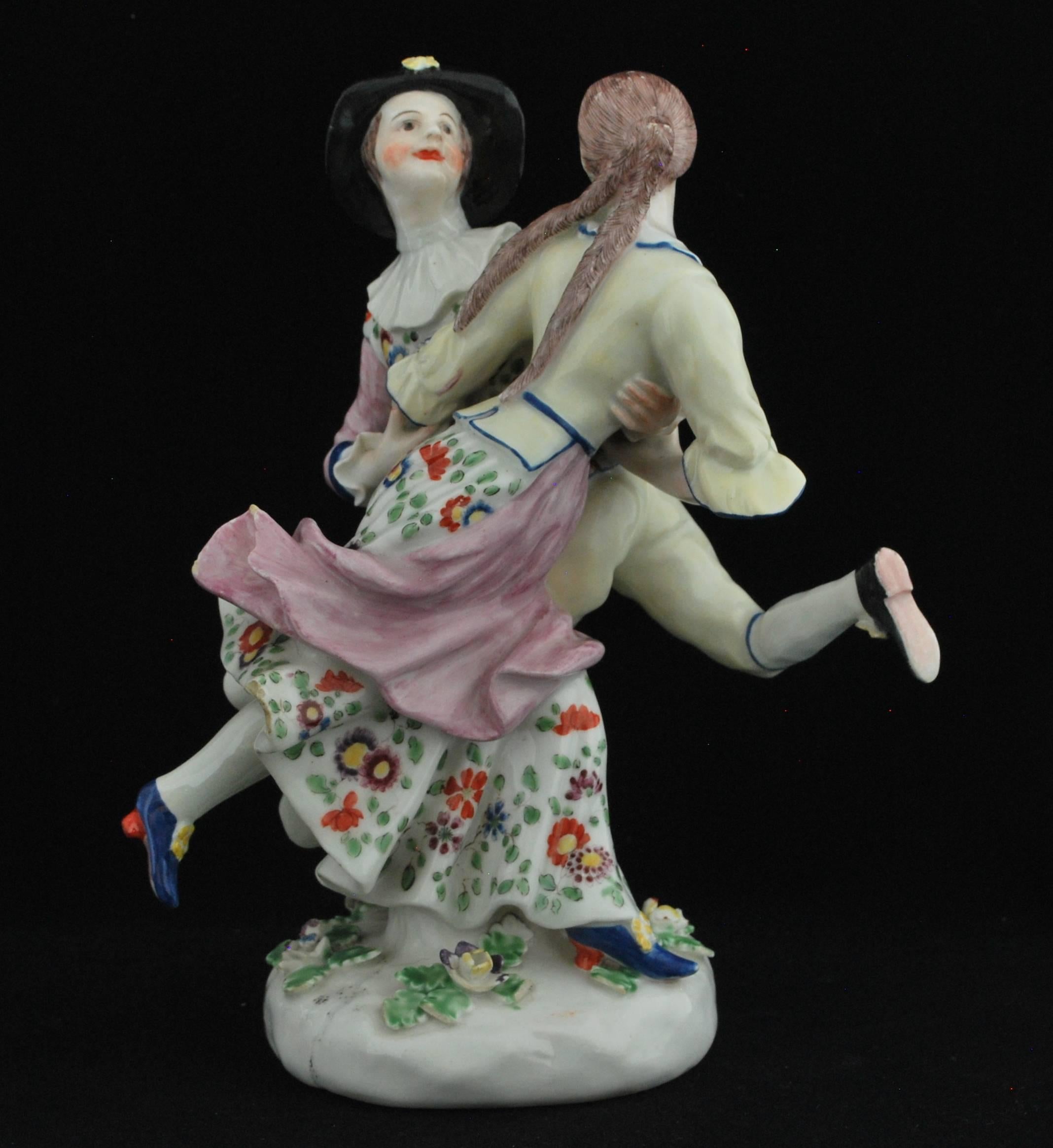 Dancers, modelled in the round, holding closely and each with one leg raised. He wears a rimmed hat with a yellow flowerhead; she bare headed with long soft brown plaits at her back. Both in costumes of pale yellow trimmed in blue, his sleeves and