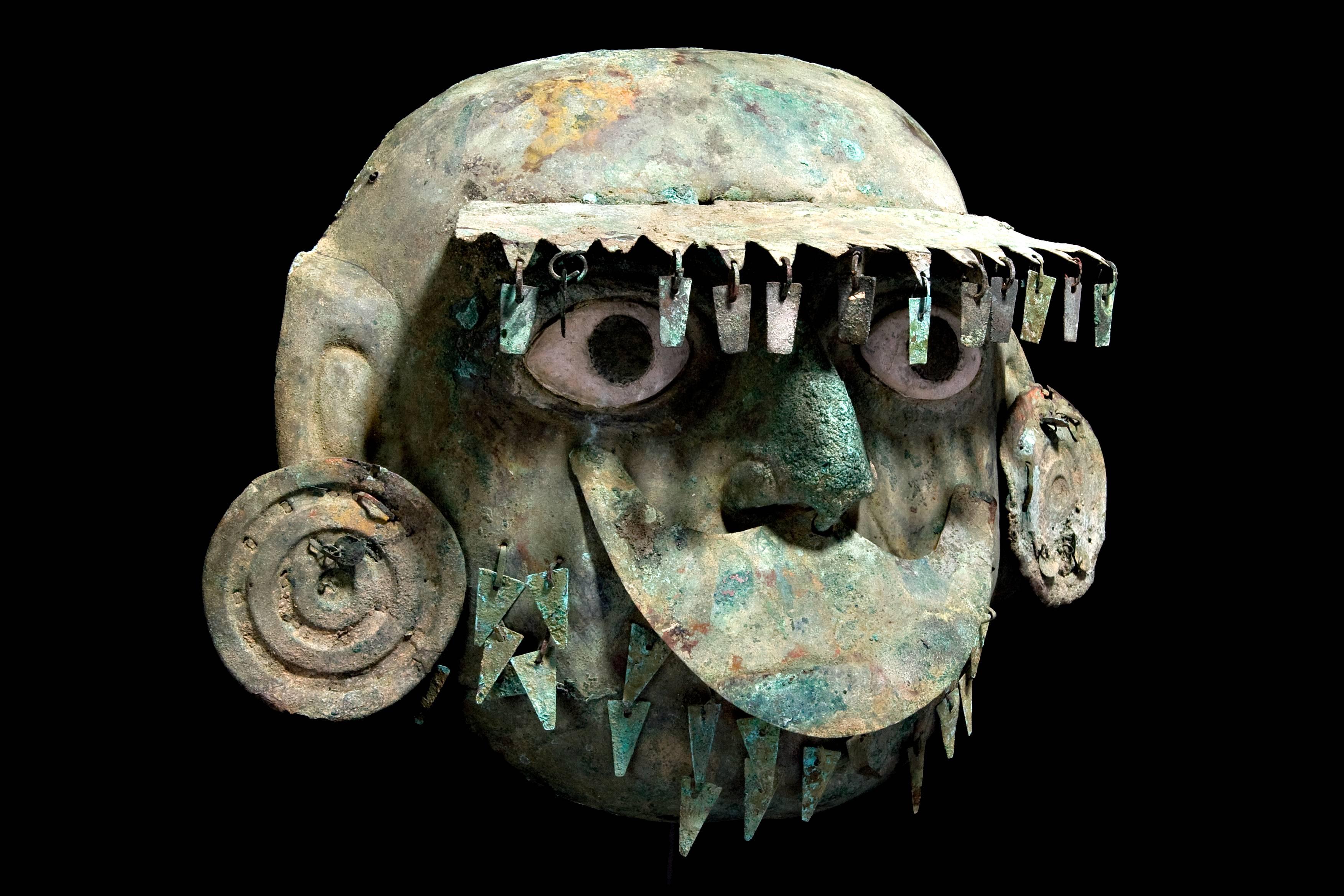A large coper mask of a dignitary with almond shaped, inset shell eyes with pupils and a wide brim type crown. Attached at the brow line, a row of danglers is suspended, aligning the brow. He is also wearing large, round ear ornaments decorated with