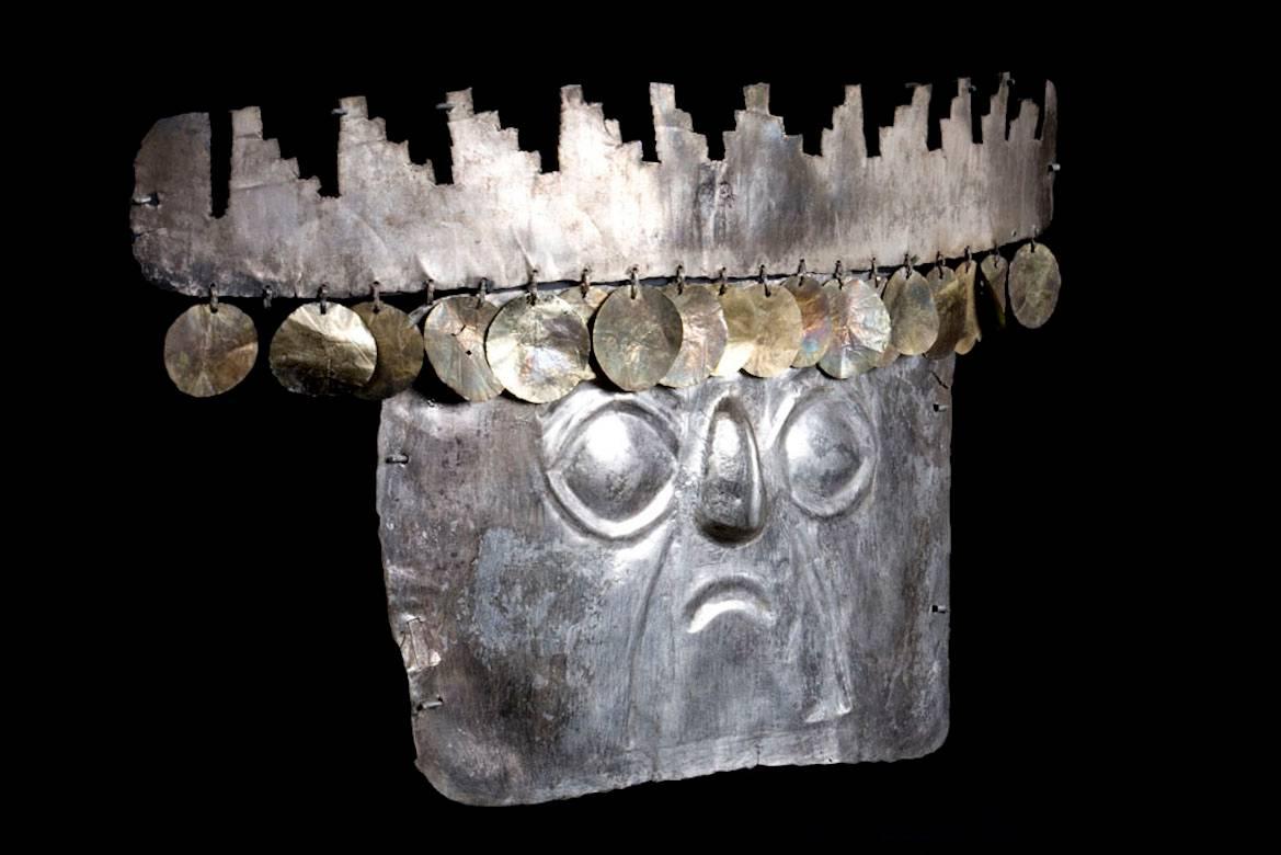 A rare, sheet silver human face mask with simple relief facial features including large, almond shaped eyes and downturned mouth. Classic stepped pyramid headdress with round, very thin sheet gold sequin adornments. Perforated along the edges for