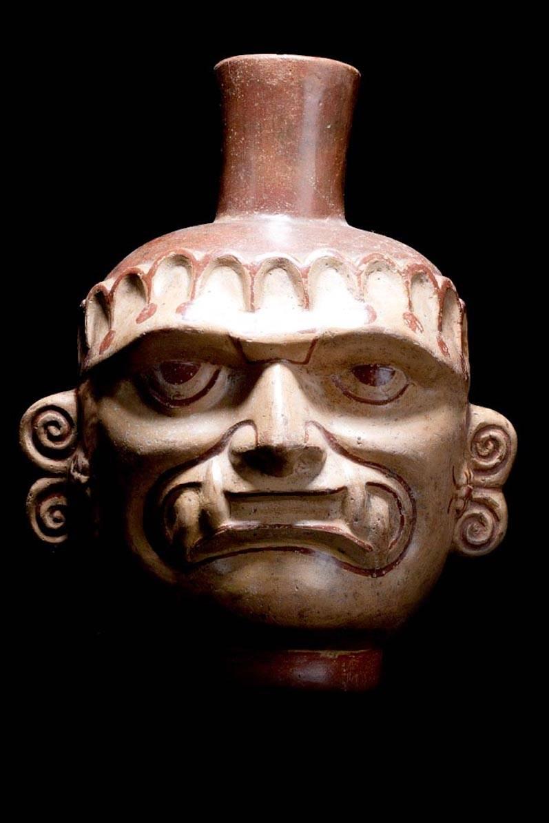 Exceptional large Mochica head pot depicting a man wearing a copper Ai Apaec jaguar teeth mask. Glazed and stone-polished portrait of a lord or shaman. Painted serpent ear ornaments with what appears to be insects. Headband with painted dangles. An