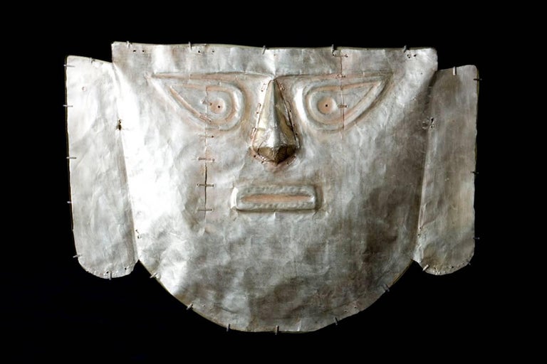 A large sheet gold mask depicting a stylized human face having an individually fashioned and applied nose. Large low relief repousse eyes and rectangular mouth. Long rectangular ears with rounded lobes having a simple beaded lobe border and pointed