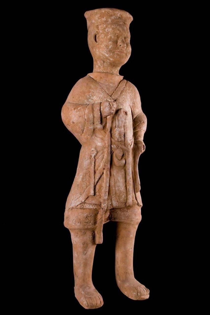 Huge hollow molded terracotta, standing male figure wearing a hip-length multi-layered robe with a collared neck. Implements suspended from his waist, holding a staff in his right hand and wearing a disc type cap. Some earthen encrustation on