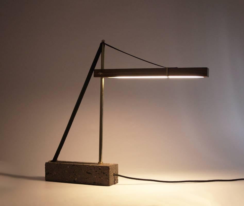 A clear homage to a construction crane's towering yet delicate balance, the Grua lamp emits warm diffuse lighting. Anchored by a block of quarry stone, a slender golden brass rod surges to sustain a walnut plank from which warm LED light showers.