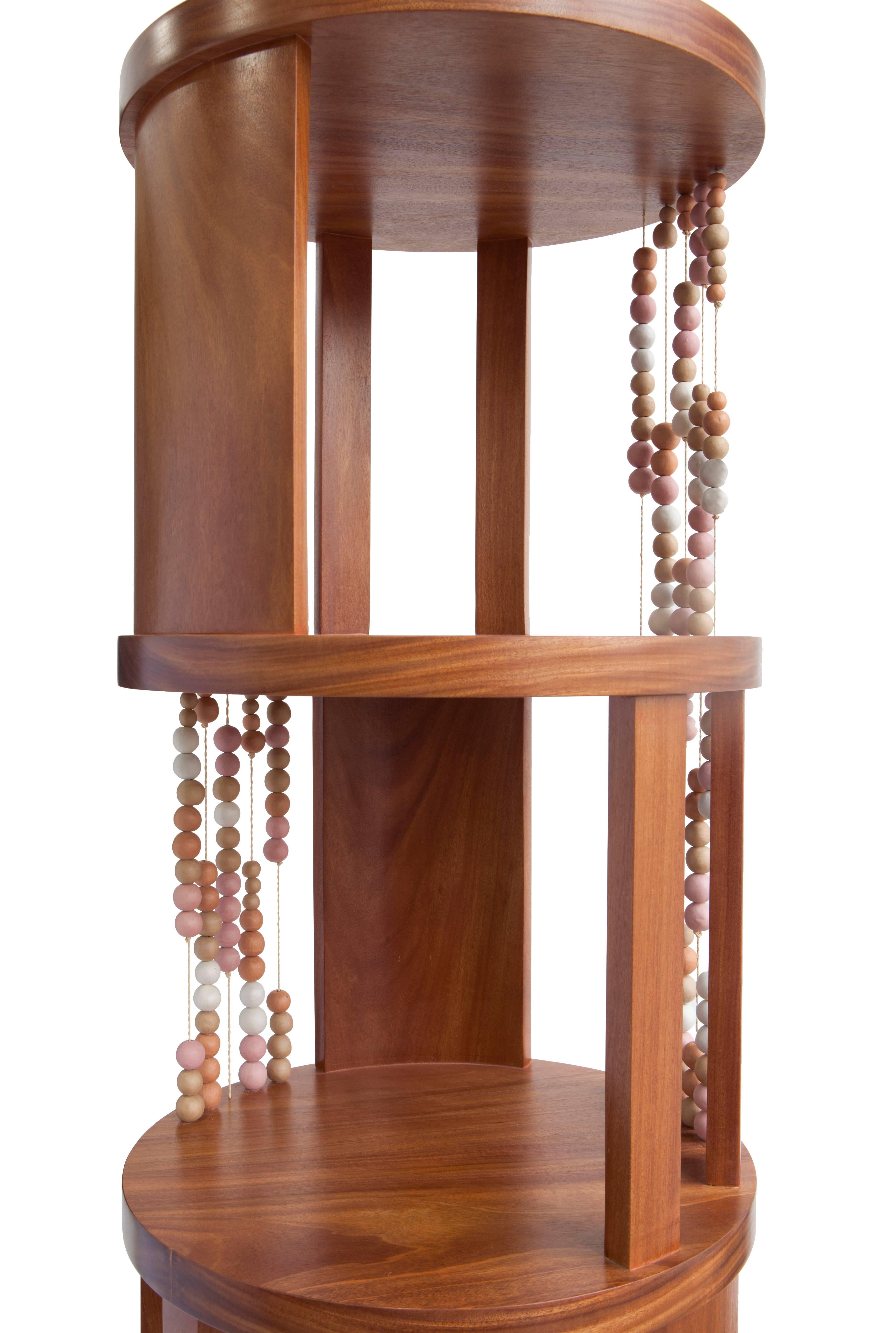 All the levels of this round bookcase, made of cabreuva hardwood, are permeated by buriti palms yarns with ceramic beads, creating different compositions depending on the angle we look at. All the soft colored shades of the ceramic come from the