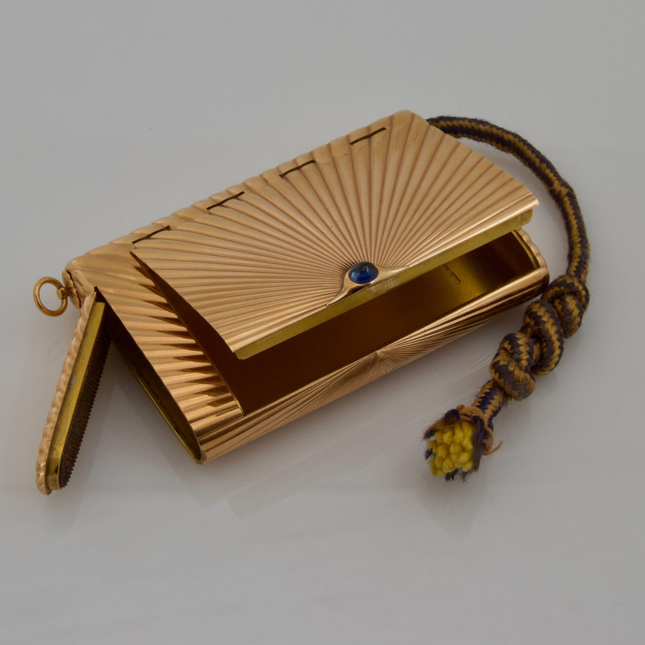 An 18-carat pink gold case with invisible hinged lid, the short ends rounded, the top and bottom engraved with a sunburst reeded pattern. The end with a vesta case compartment. Violet and yellow tinder cord retained by a ring. Cabochon sapphire