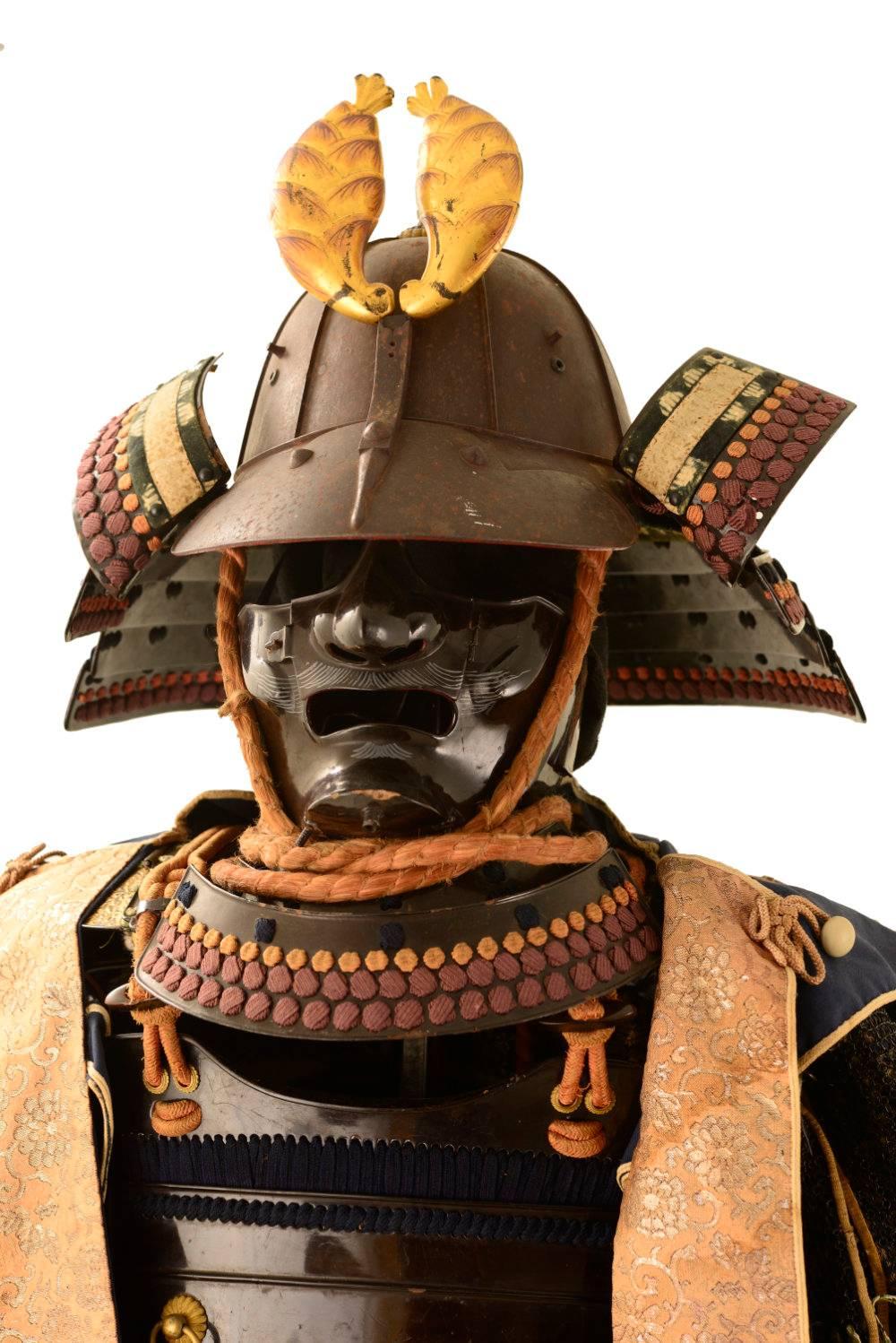 The heavy iron ridged eight plate helmet, attributed to famed smith Myochin Munesuke, with an elegantly flaring neck guard is surmounted by the apricot blossom clan symbol of the Nabeshima clan.  A smooth faced mask, and red madder silk cross knots