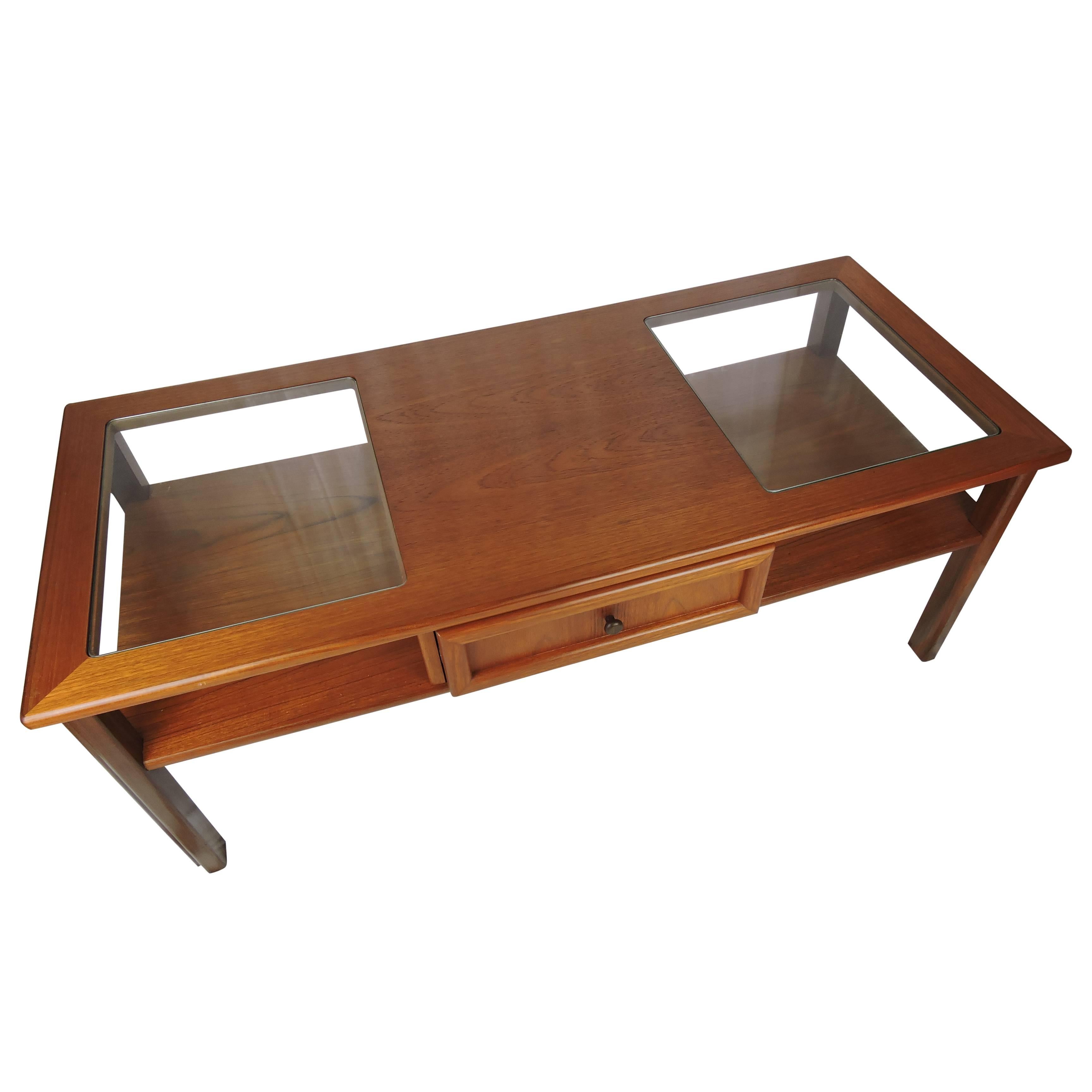 A British G-Plan teak low coffee table with a single central frieze drawer opening at both ends. It features a glass topped section over shelves at either end. Table stands on shaped legs.