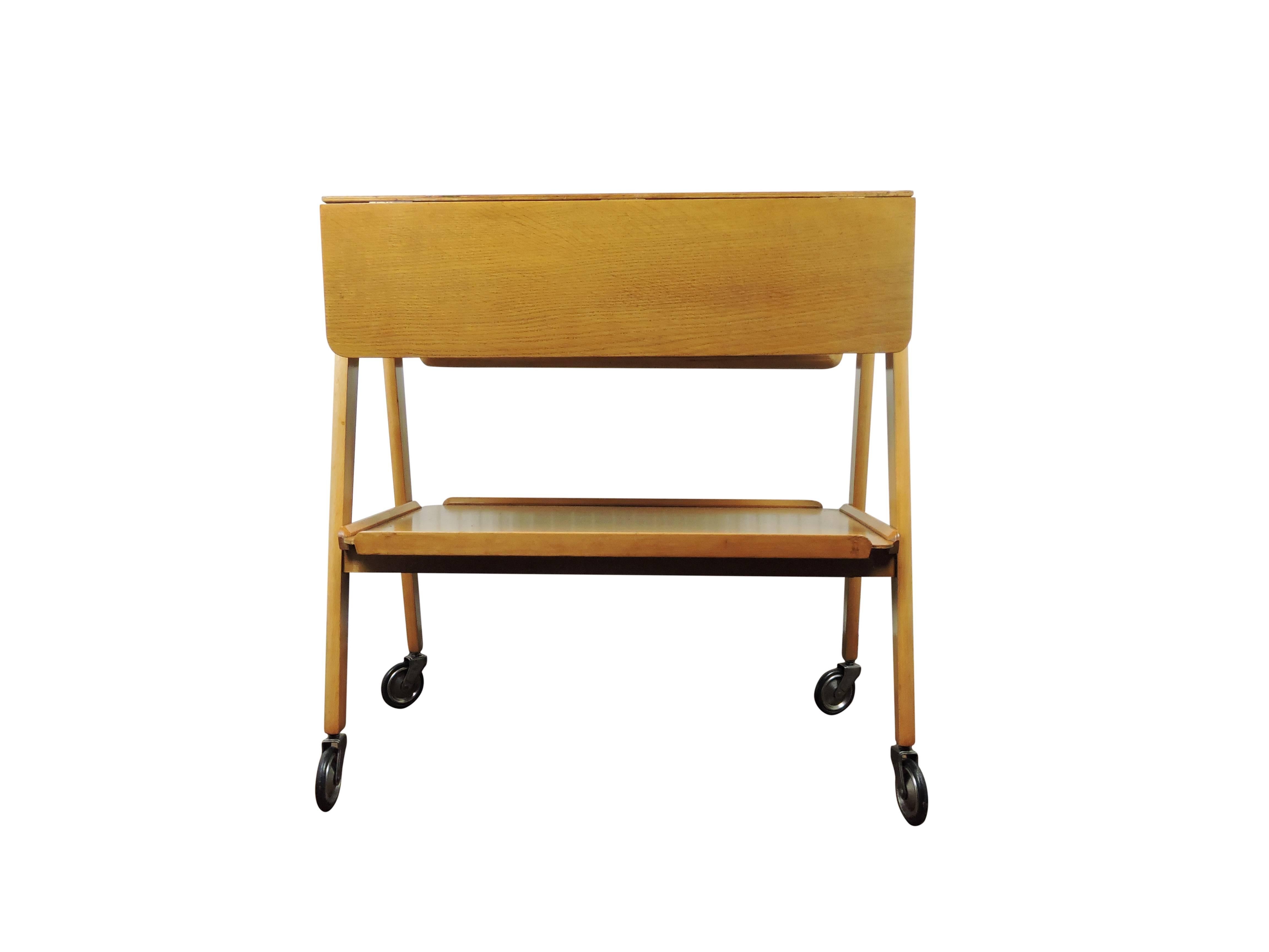 An English blonde oak Remploy two-tier trolley. The trolley features two hinged flaps which open up to produce a table top. The trolley is on large castor wheels for easy moving.