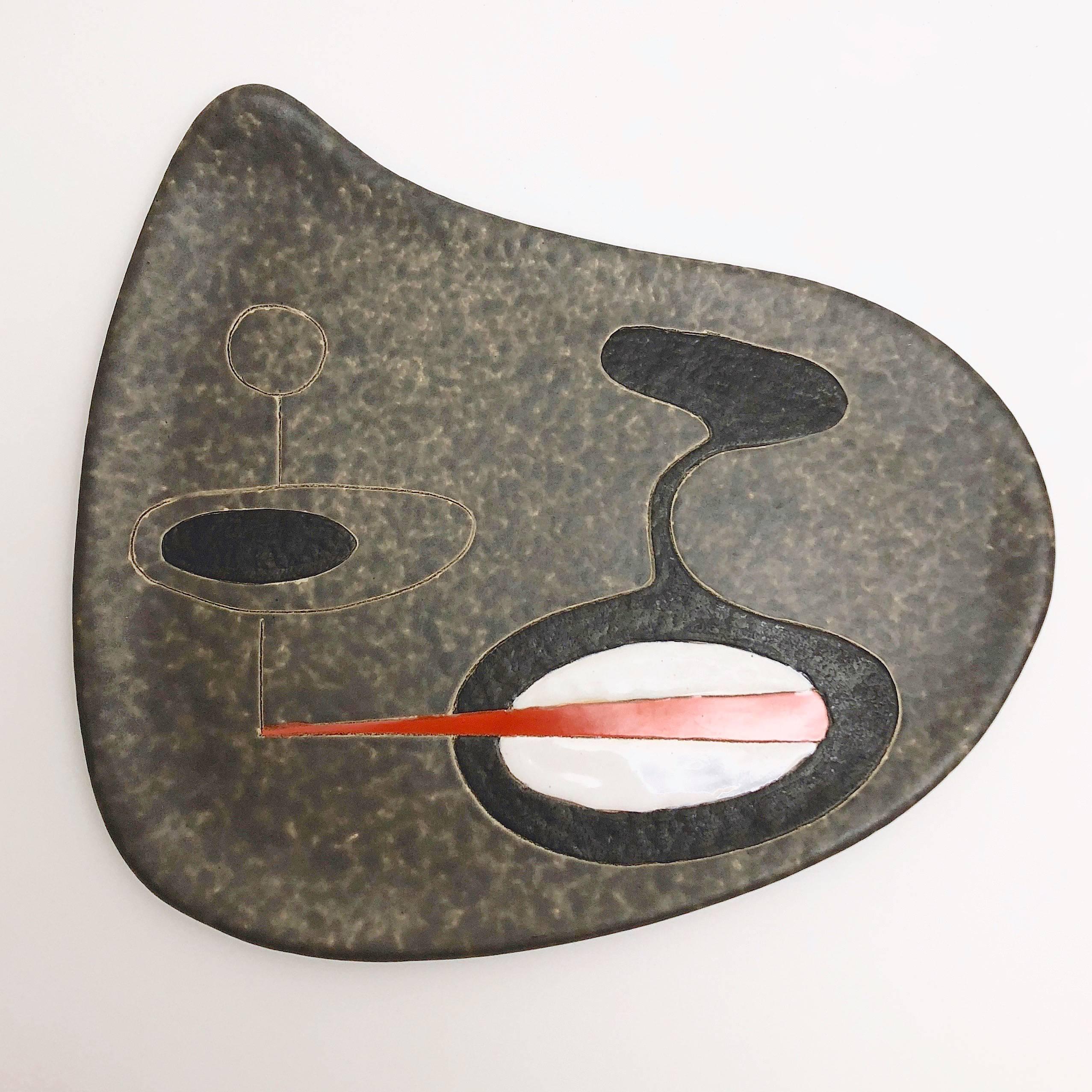 An organic shaped earthenware dish glazed in cloudy warm grey, with abstract and geometric decoration engraved and over-glazed in black, white, and orange. 

Typical Mid-Century Modern abstract and geometric design, reminiscent of Joan Miro´ or