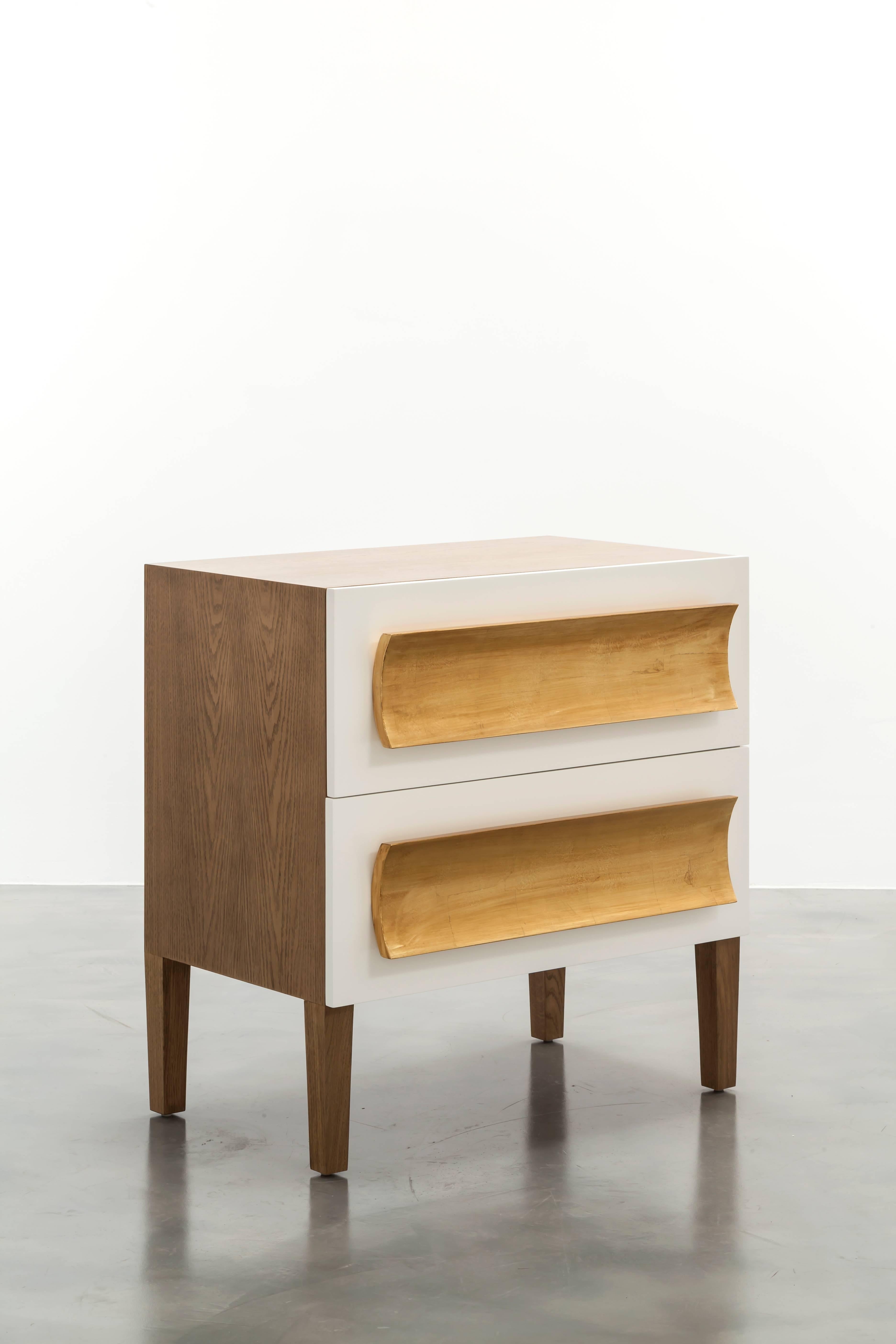 The Recaimer nightstand features a unique mixed finish design with an oak body, lacquer drawer fronts and gold leaf hand-carved pull details. Drawers are standard soft close with hidden glides. Showroom samples sold as is. Natural walnut with white