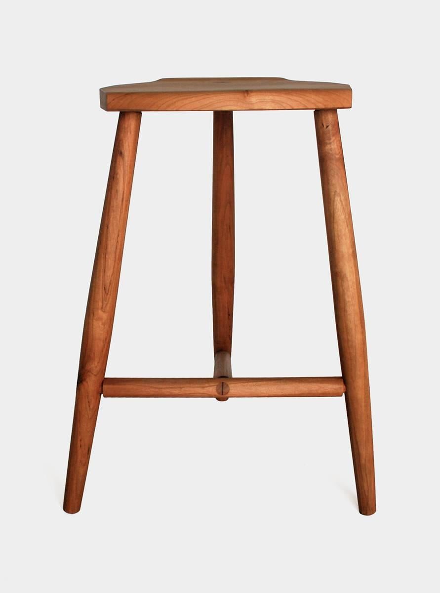 New York Heartwoods' black cherry solid wood counter height Jordan Stool is influenced by Shaker and Mid-Century design; created to be comfortable, light and easy to move; and features three turned legs, a unique hand-carved faceted seat, and wedged