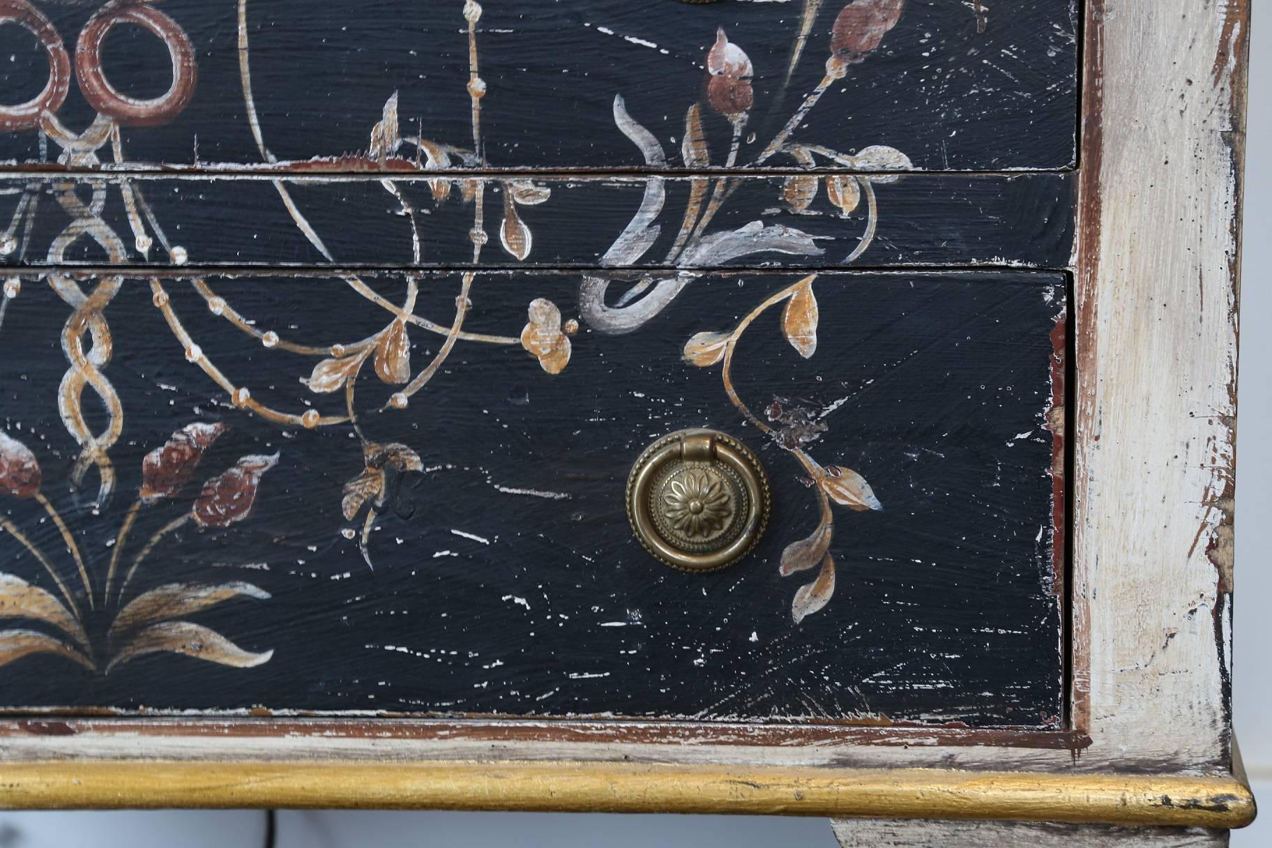 Hand-painted three-drawer chest in distressed ivory, black and gold colors with urn and floral decoration on front, top and sides. Brass accents, Italy.