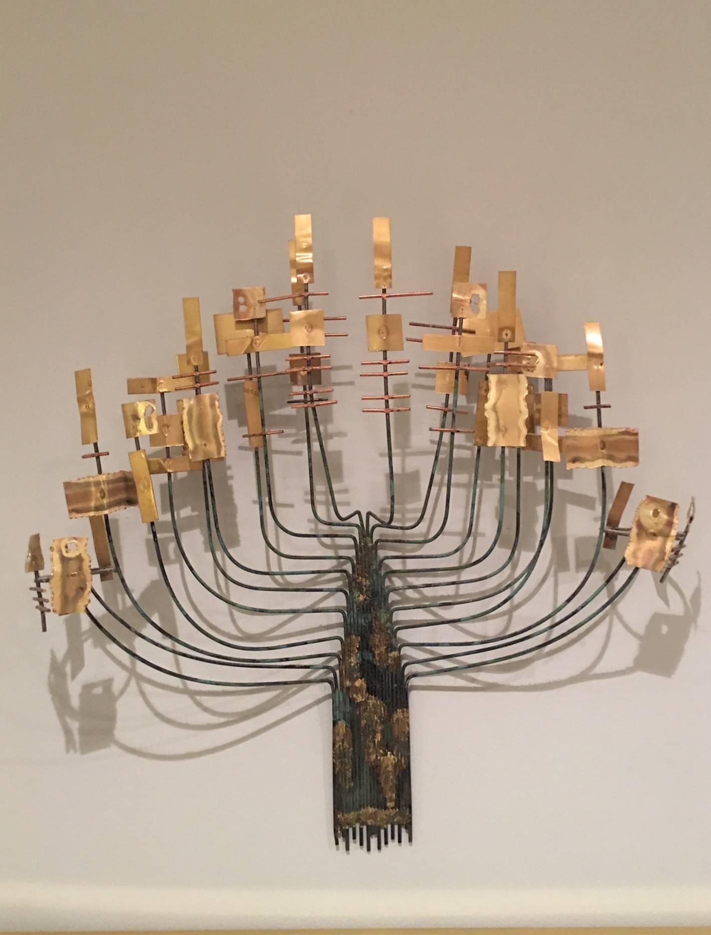 This lovely wall sculpture is in the manner of Curtis Jere, but is unsigned. It is crafted of brass and metal with tones of gold and Verdigris. Reminiscent of a menorah, it is the perfect small accent piece in a photo grouping or above a small