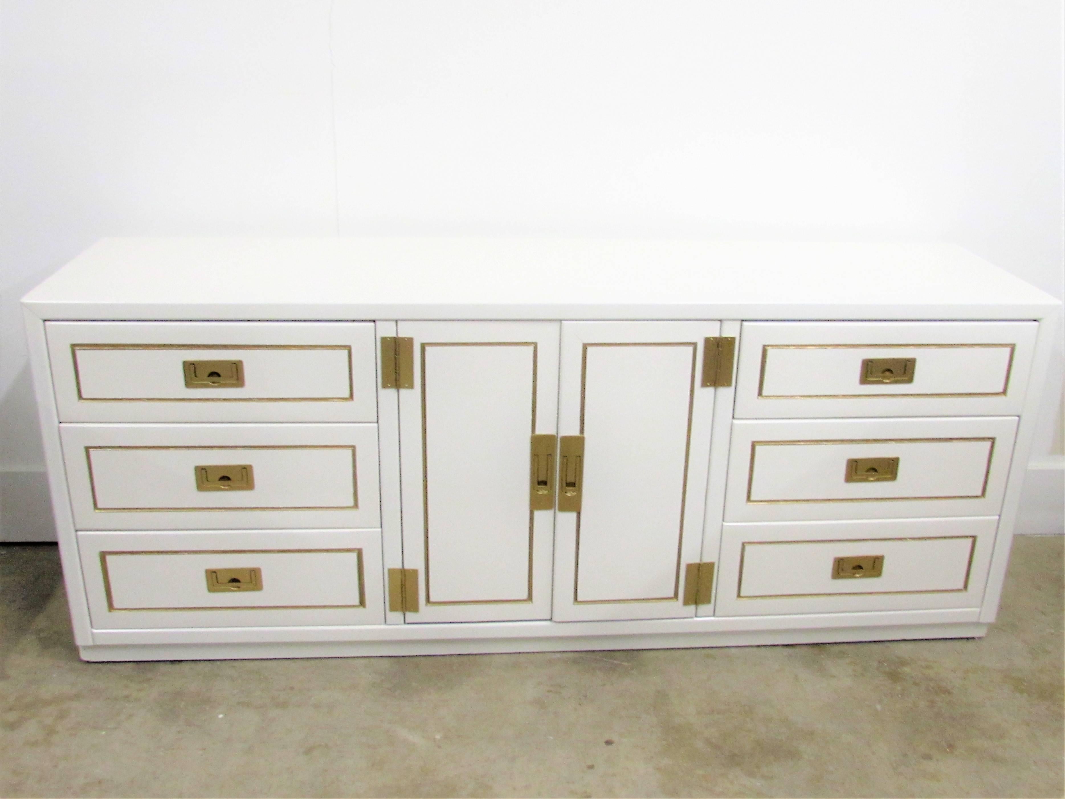 Nine-drawer lacquered campaign credenza in alabaster white gloss finish with gilded accents and recessed campaign hardware in our custom gold under brass. Three dovetail drawers are on each side of two swinging doors that open to three additional