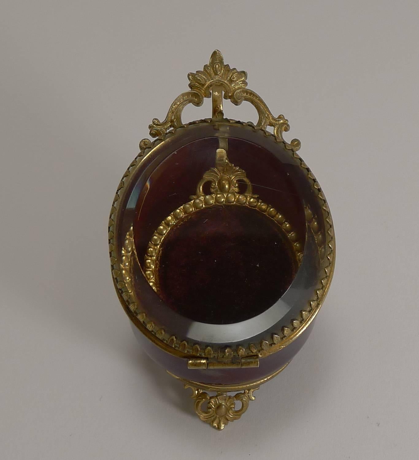 A pretty antique French pocket watch box, made from a deep red glass and lined with a rich Burgundy velvet with an original thick bevelled glass lid.

The fittings including the three pierced or reticulated feet are made from gilded brass. Once