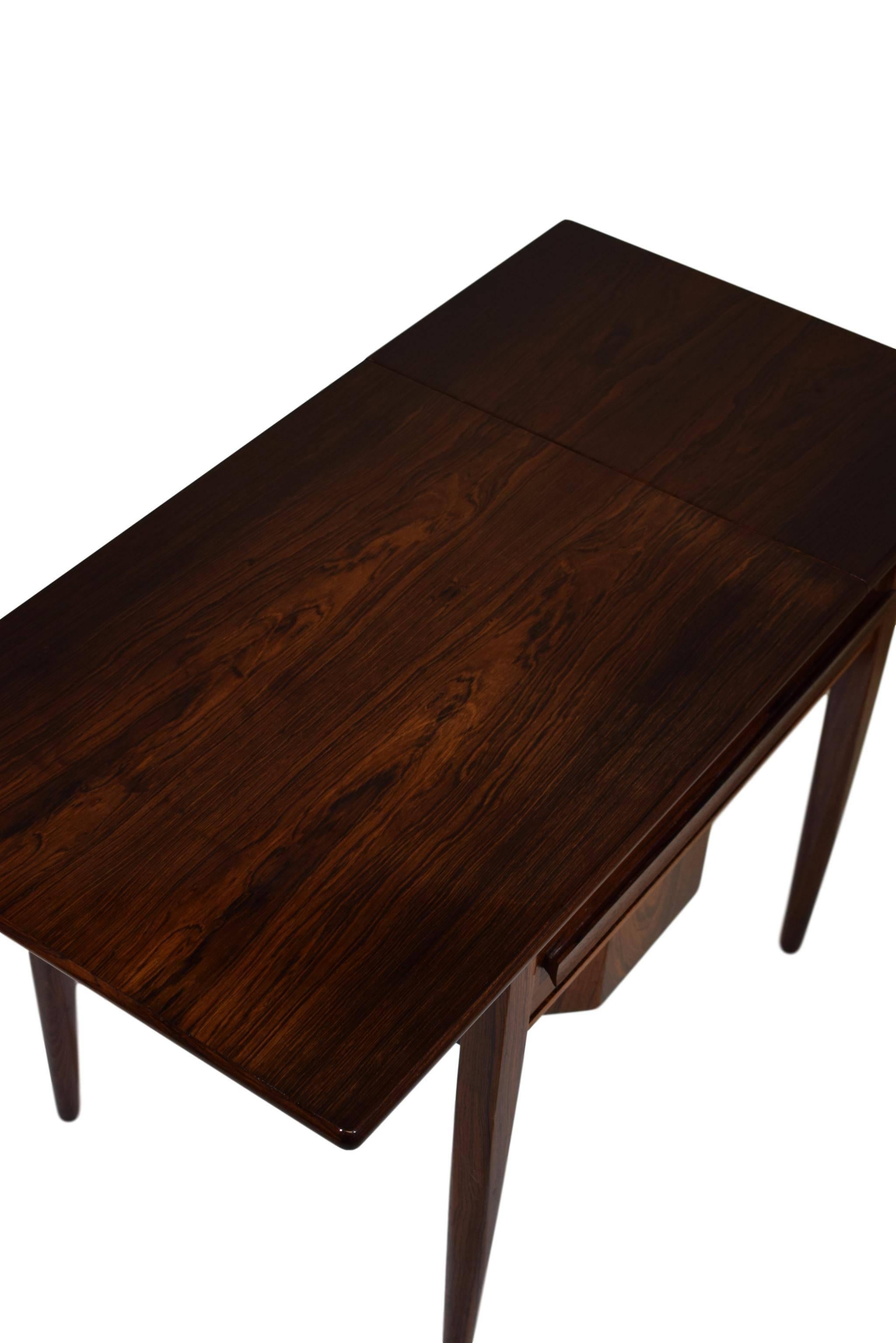 A Danish midcentury sewing table with drop leaf and one drawer for yarns. By Johannes Andersen. Rosewood veneer and tapered solid rosewood legs.
 