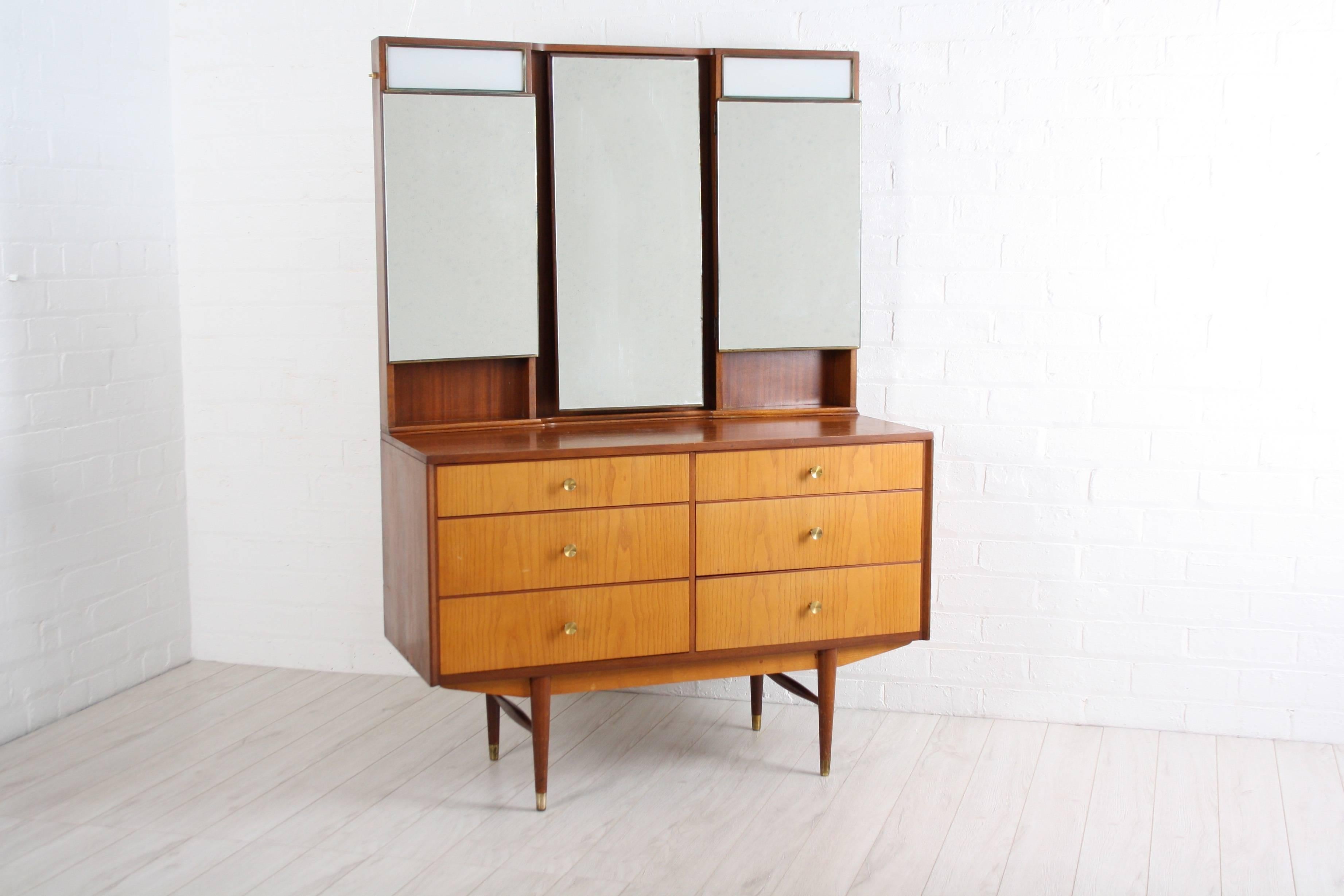 A beautifully sleek 1960s teak dressing table with a triple backlit mirror over an arrangement of six drawers on tapering legs. 

Handy hidden shelving behind the right and left mirrors and the light is in excellent working order with new wiring.