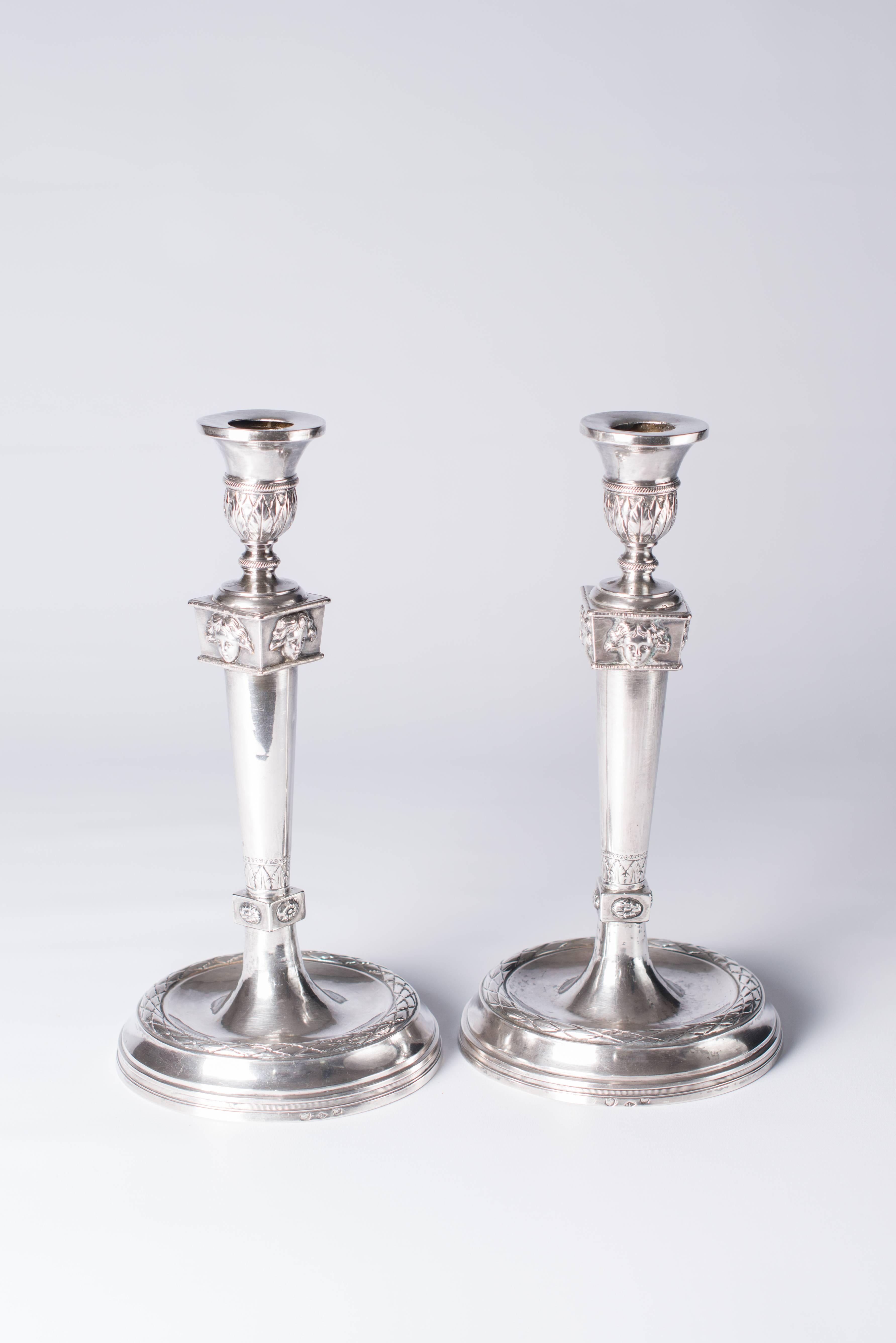 Pair of French Empire silver candlesticks are well casted. Each on a circular moulded base with sunken centre, the plain tapering stem between a small and larger square knob with stylised foliage and masks, the vase-shaped candleholder with acanthus
