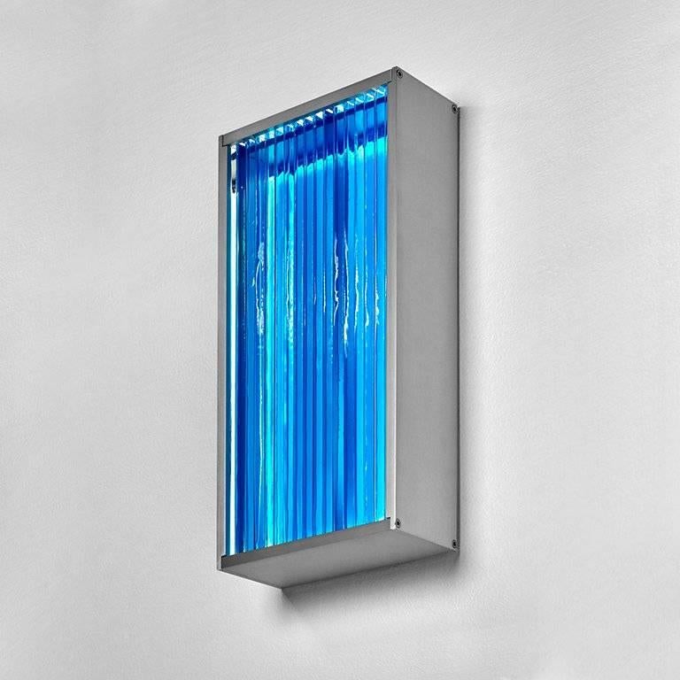 This wall sconce is created by laminating clear glass pieces together in a vertical pattern using pigmented adhesive. An LED light panel illuminates the glass and transparent pigment from behind. In a sense, the glass acts as a lens, concentrating