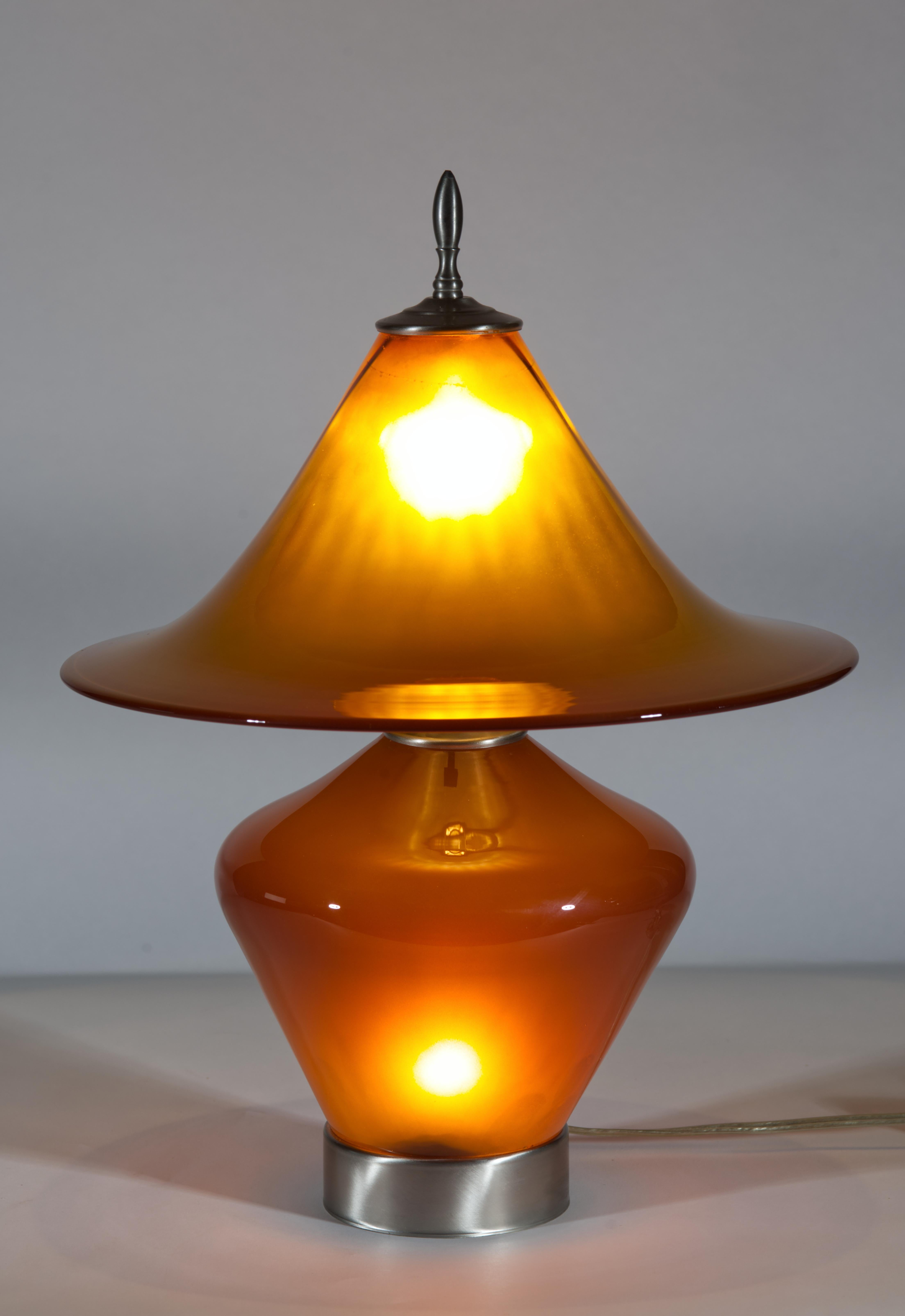 Vintage Steven Correia art glass table lamp features an elegant, unusual shape and a striking deep amber glass base and shade. The design shows a strong influence of Streamline Modern and Art Deco styles on Post Modern aesthetics. Silver colored