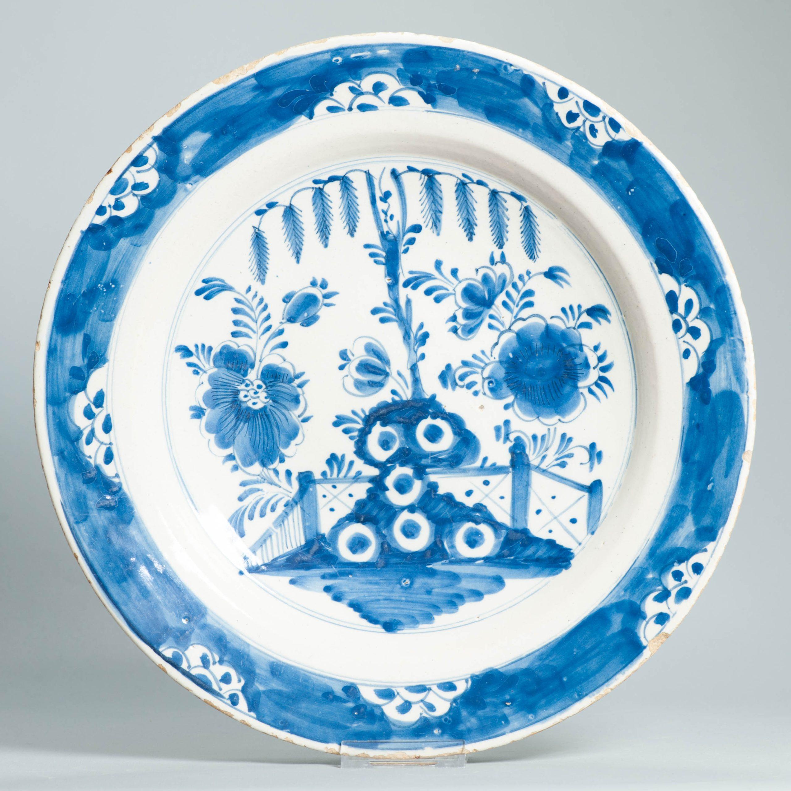 Un piatto blu e bianco di Delft olandese del XVIII secolo, affascinante e di dimensioni generose, ispirato ai disegni delle porcellane cinesi del periodo Kangxi. La scena centrale è caratterizzata da un giardino splendidamente stilizzato con piante