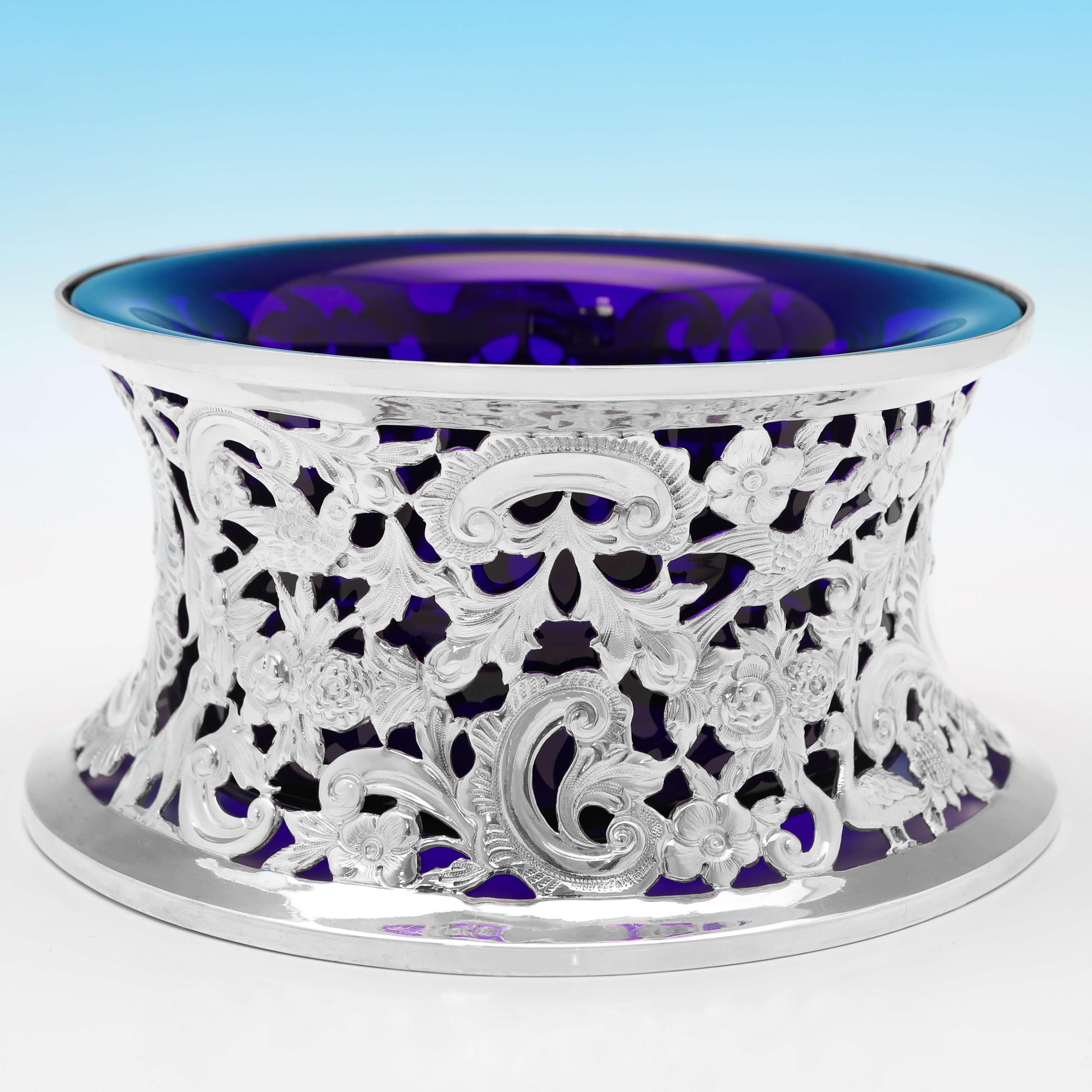 Con marchio a Dublino nel 1895 da Charles Langley, questo antico e affascinante anello per piatti in argento sterling presenta decorazioni cesellate e traforate che raffigurano fiori, foglie, uccelli, cani e altri animali e un rivestimento in vetro