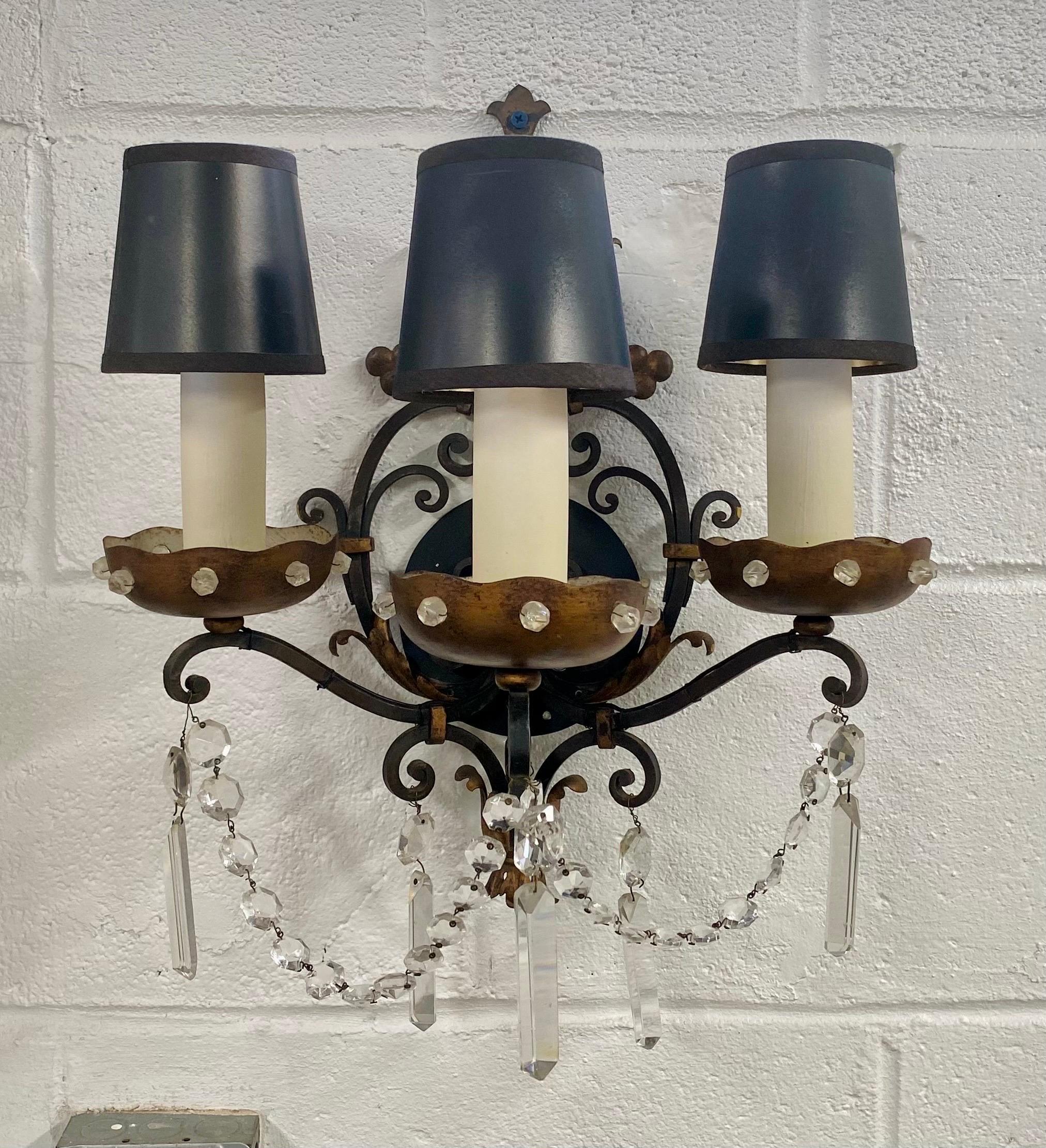This elegant pair of Maison Jansen-style sconces features wrought iron backings, each embellished with a tole bobeches boasting crystal button detail. Adorned with a combination of crystal chains and prism, the sconces each feature three arms and