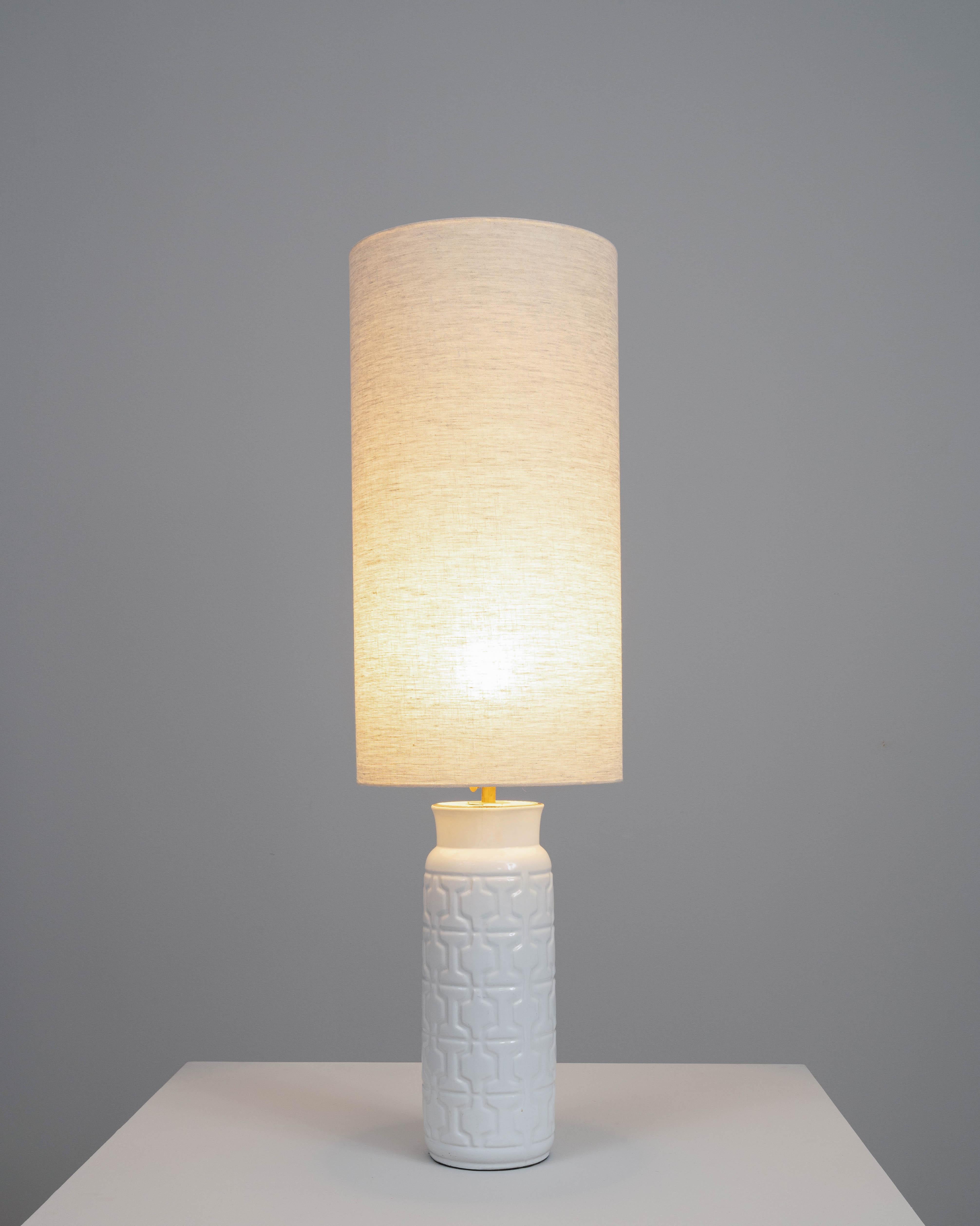Add a touch of understated elegance to your home with this early 20th-century British ceramic table lamp. The lamp features a sleek, cylindrical base with a subtle geometric pattern, beautifully glazed in a pristine white finish. Topped with a tall,