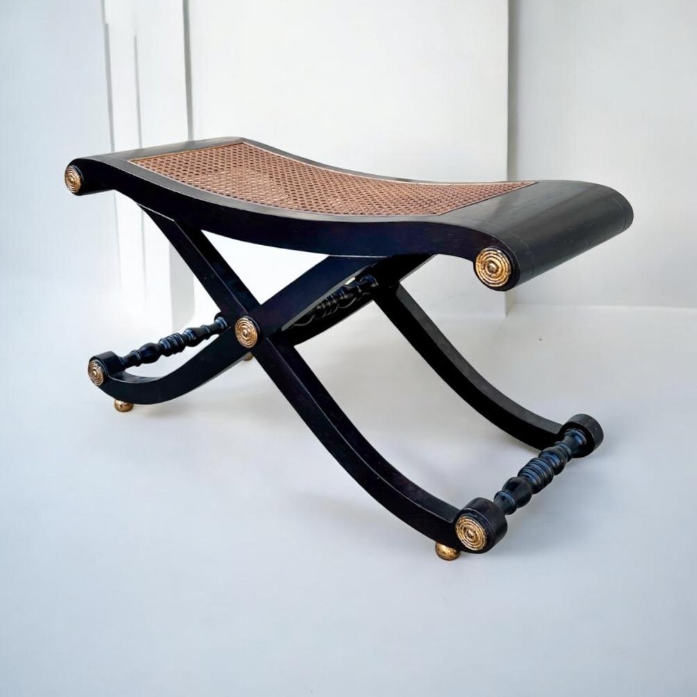 This is an ebonized x-base caned bench with gilt accents by Baker Furniture. It has timeless appeal and is in very good condition. It is marked. 