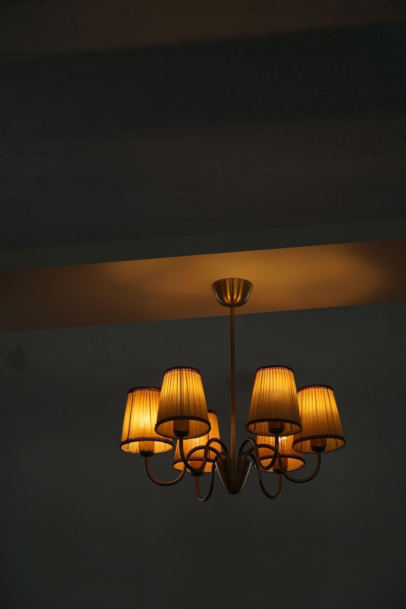 Questo lampadario svedese del Mid-Century Modern degli anni '50 presenta cinque bracci in ottone elegantemente curvati, ciascuno dotato di un paralume in tessuto morbidamente affusolato. La sua silhouette minimalista e le sue proporzioni raffinate