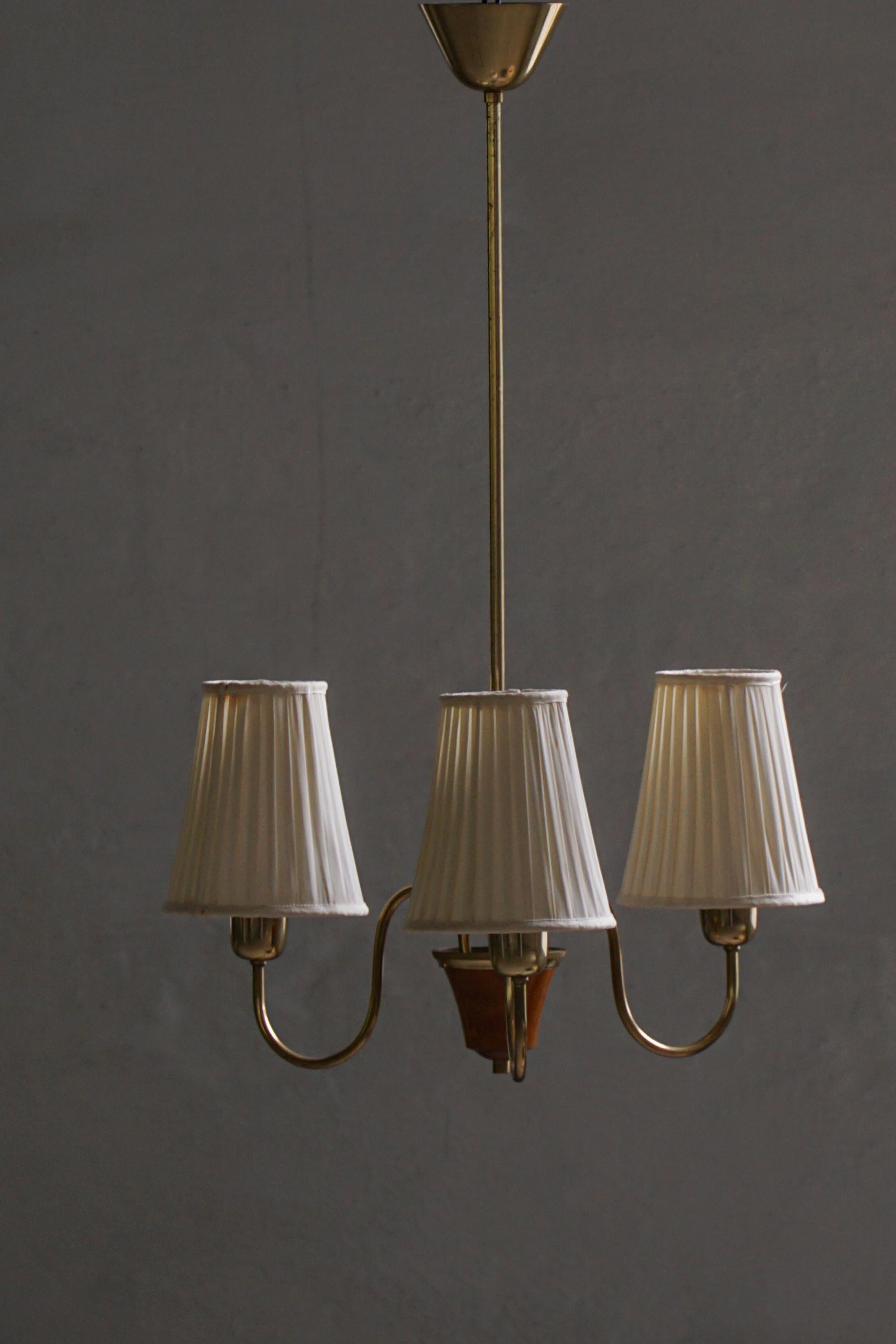 Un affascinante lampadario svedese degli anni '40 che unisce calore, artigianalità e raffinata semplicità. Il design è caratterizzato da una struttura in ottone patinato con dettagli in legno tornito, che sostiene tre paralumi in seta plissettata