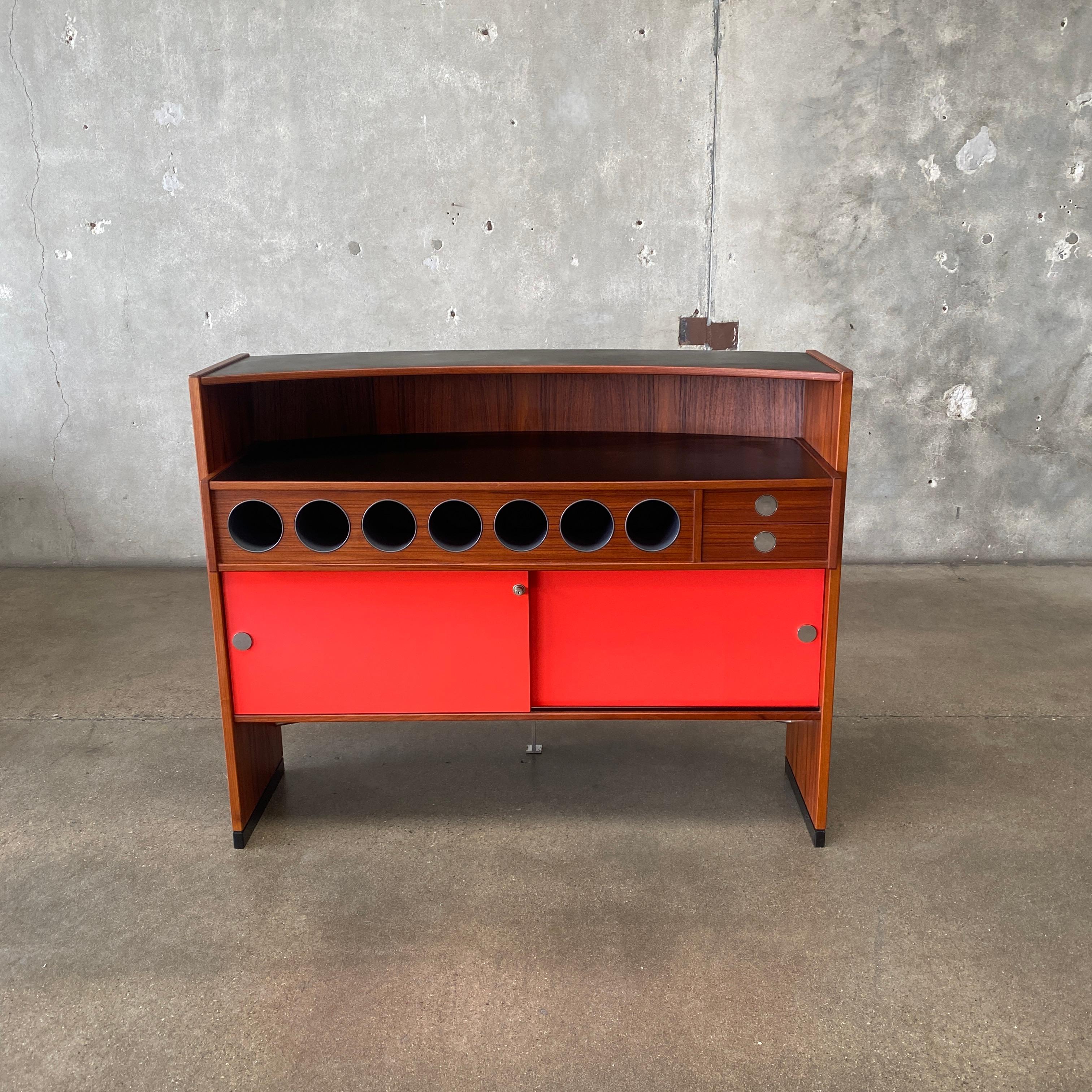 This is an iconic mid-century modern teak dry bar designed by Erik Buch for Dyrlund, made in Denmark circa the 1960s. The bar is a curved form with a chrome footrest and water-resistant black table tops. The brilliant coral sliding doors with