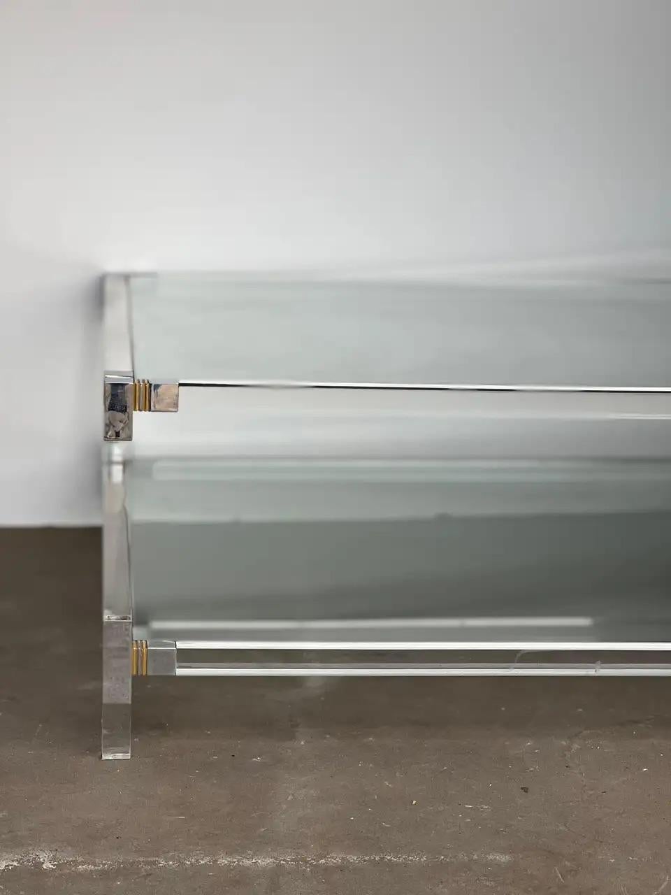 Here is a transitional imported 1970s Mid-Century Modern brass, chrome, glass and lucite Italian coffee table from Italy. The coffee table features two shelves of glass with lucite tubing around the edges that each end into chrome and brass