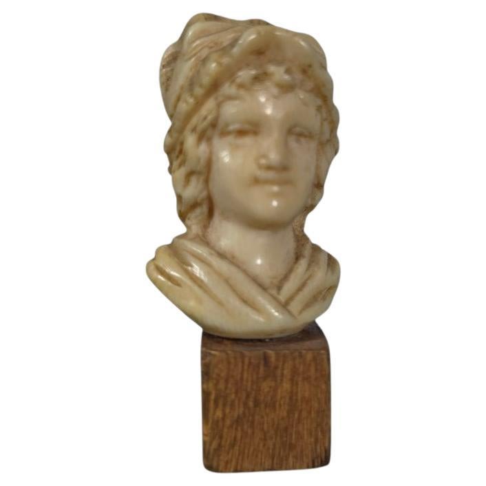 Delicate, finely carved bone bust depicting a young woman dressed in 19th-century fashion. The face is gently rendered, revealing a serene and refined expression. The piece is mounted on a simple wooden base, highlighting the finesse of the