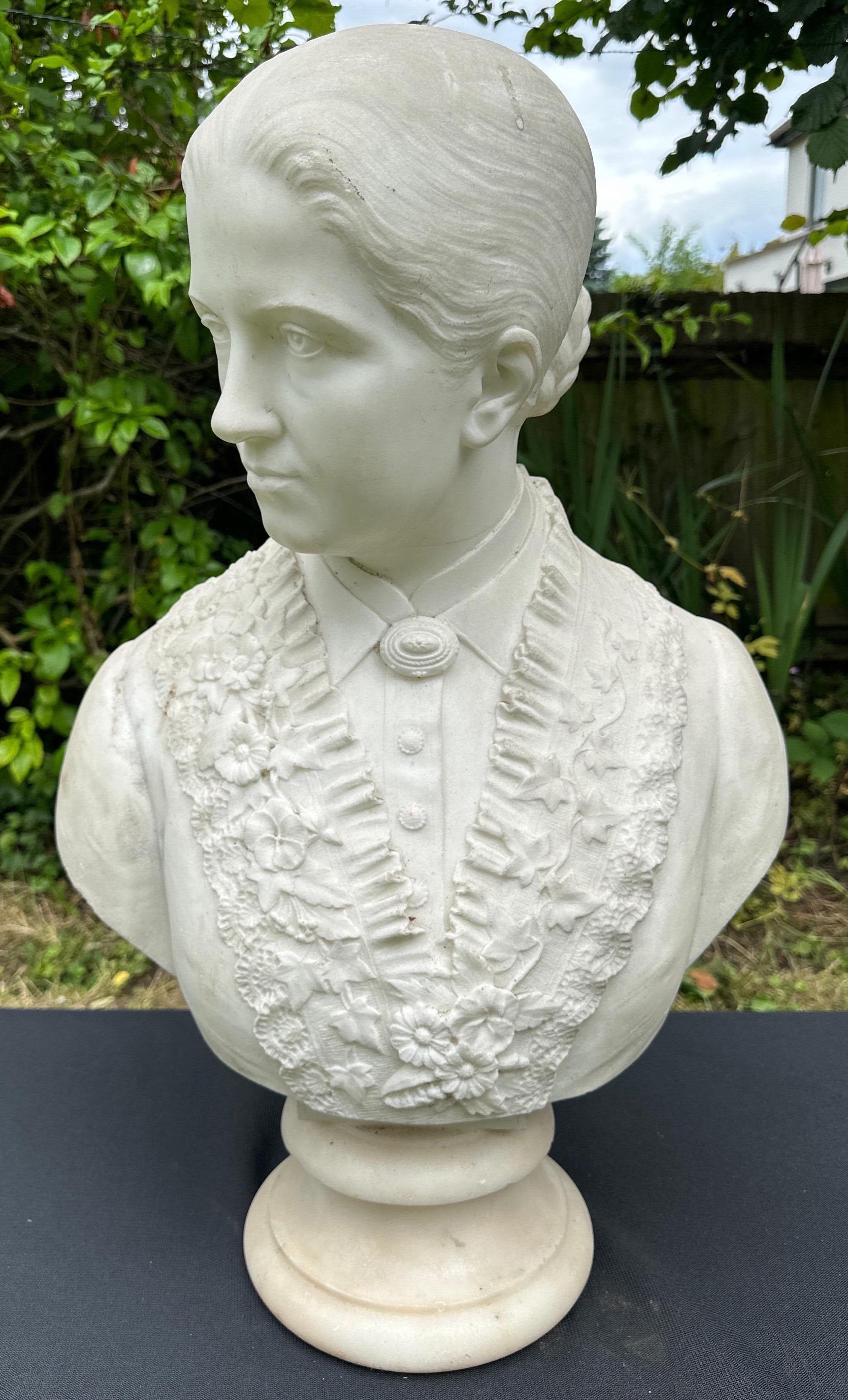 A white marble bust of an elegant lady with her hair in a bun. Wearing a floral embroidered flowery dress. 
Sitting on a white marble circular base.