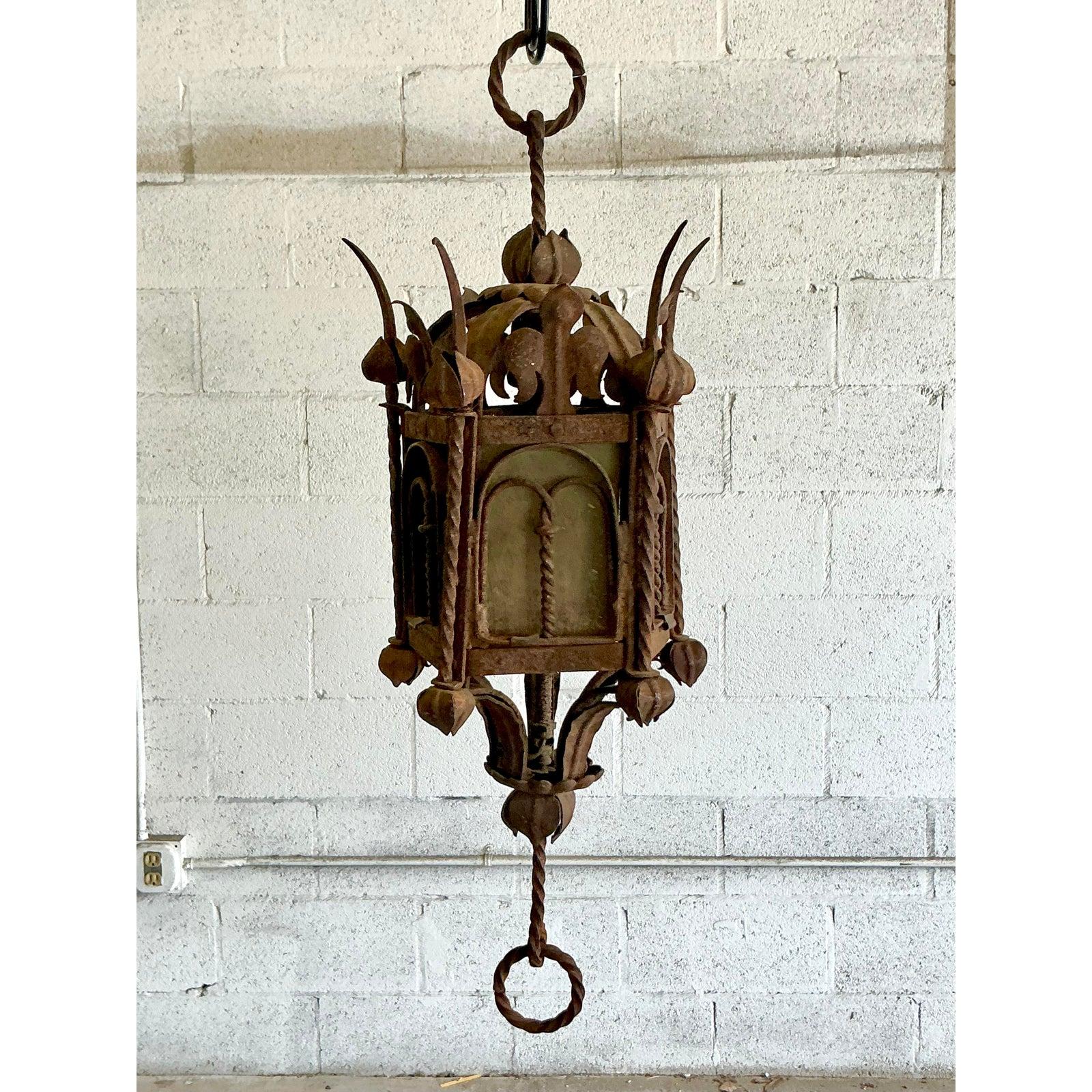 Evoking the old-world romance of Spanish Revival architecture, this stunning vintage lantern is a testament to rugged, artisanal craftsmanship. The heavily patinated wrought iron frame features intricate, Gothic-inspired details and a beautifully
