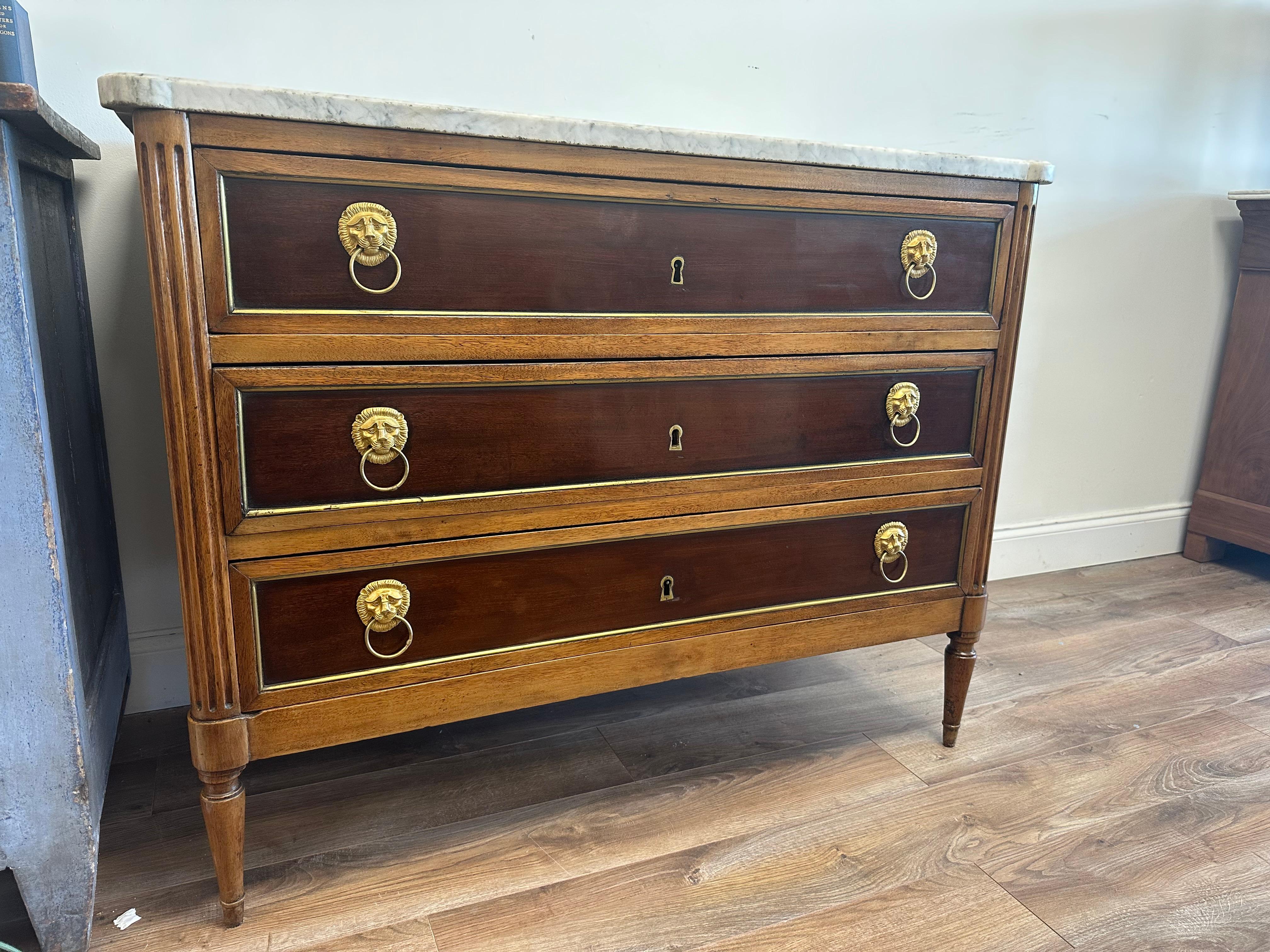 This Louis XVI Chest of Drawers has 3 drawers with a marble top. The hardware is brass lion carved rosettes with a drop ring pull. The finish has 2 stain colors (a darker color paired with a lighter walnut color) for a two-tone stained finish. The