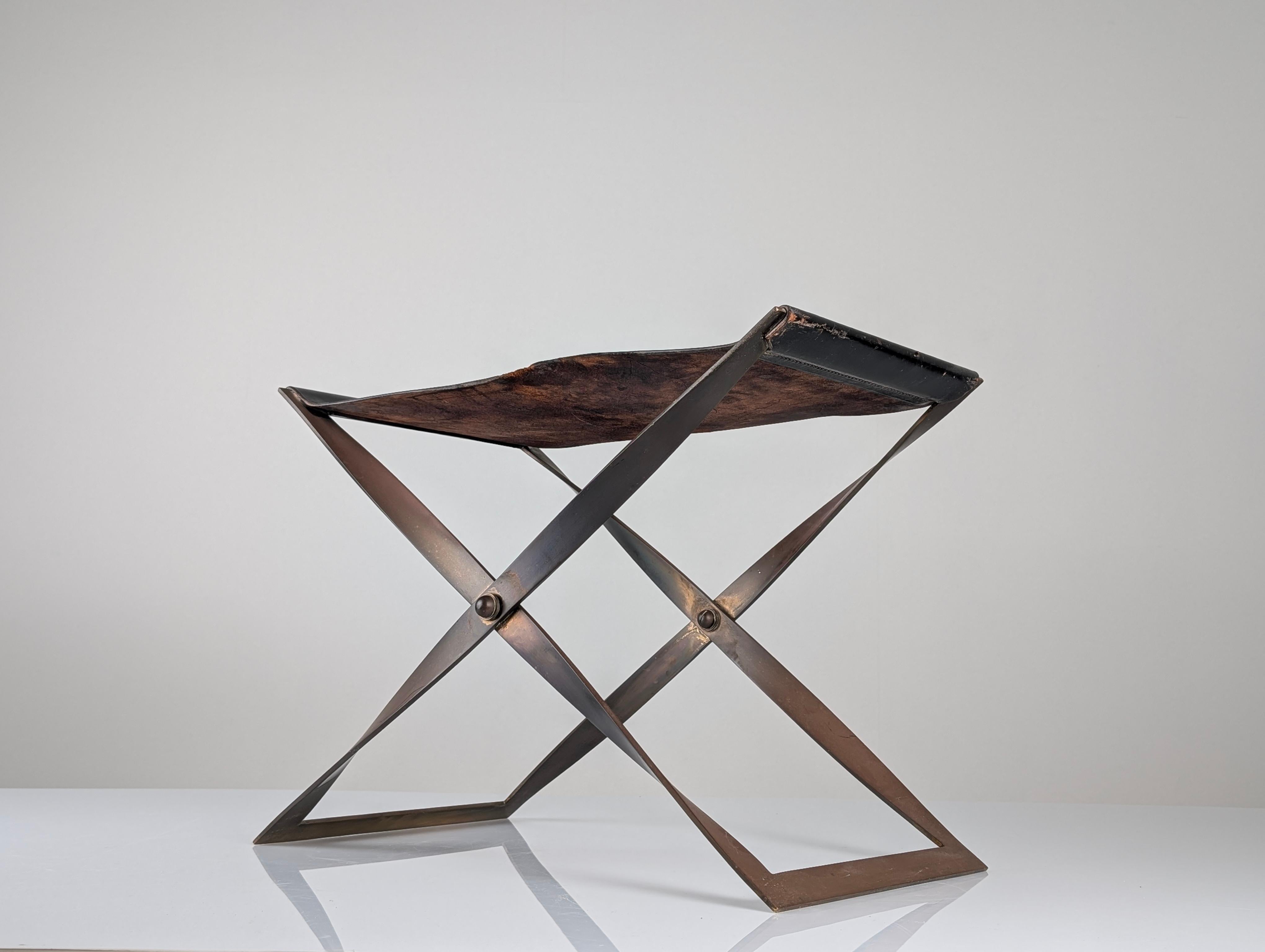 Sgabello danese pieghevole degli anni '60 con struttura in acciaio spazzolato e seduta in pelle marrone scuro con patina originale. La sua silhouette a X e la seduta sospesa definiscono un'estetica pulita, funzionale e scultorea, tipica del design
