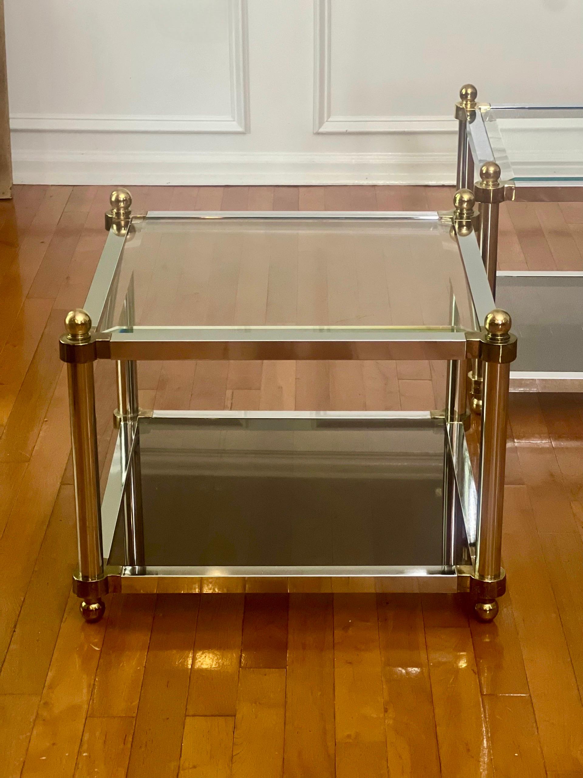 Hübsches Paar zweistöckiger quadratischer Glastische aus Messing und poliertem verchromtem Stahl aus den 1970er Jahren in der Art von Maison Jansen.

Die untere Ebene besteht aus Rauchglas und die obere aus transparentem, abgeschrägtem Glas. Oben