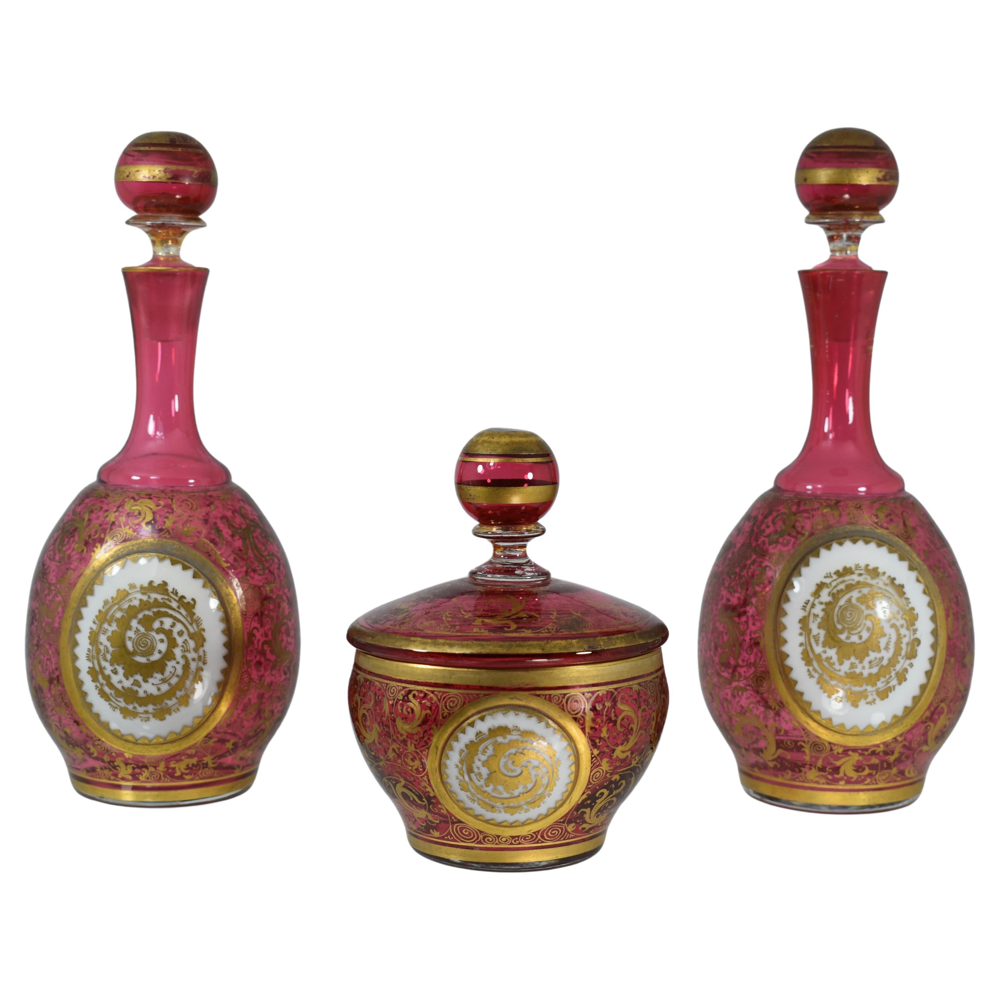 A Set of two similar bottles with stoppers and one bowl with cover made of cranberry glass with white opaque overlay, the bottles with circular baluster formed body, richly decorated with fine gilding all around, all three parts are applied with