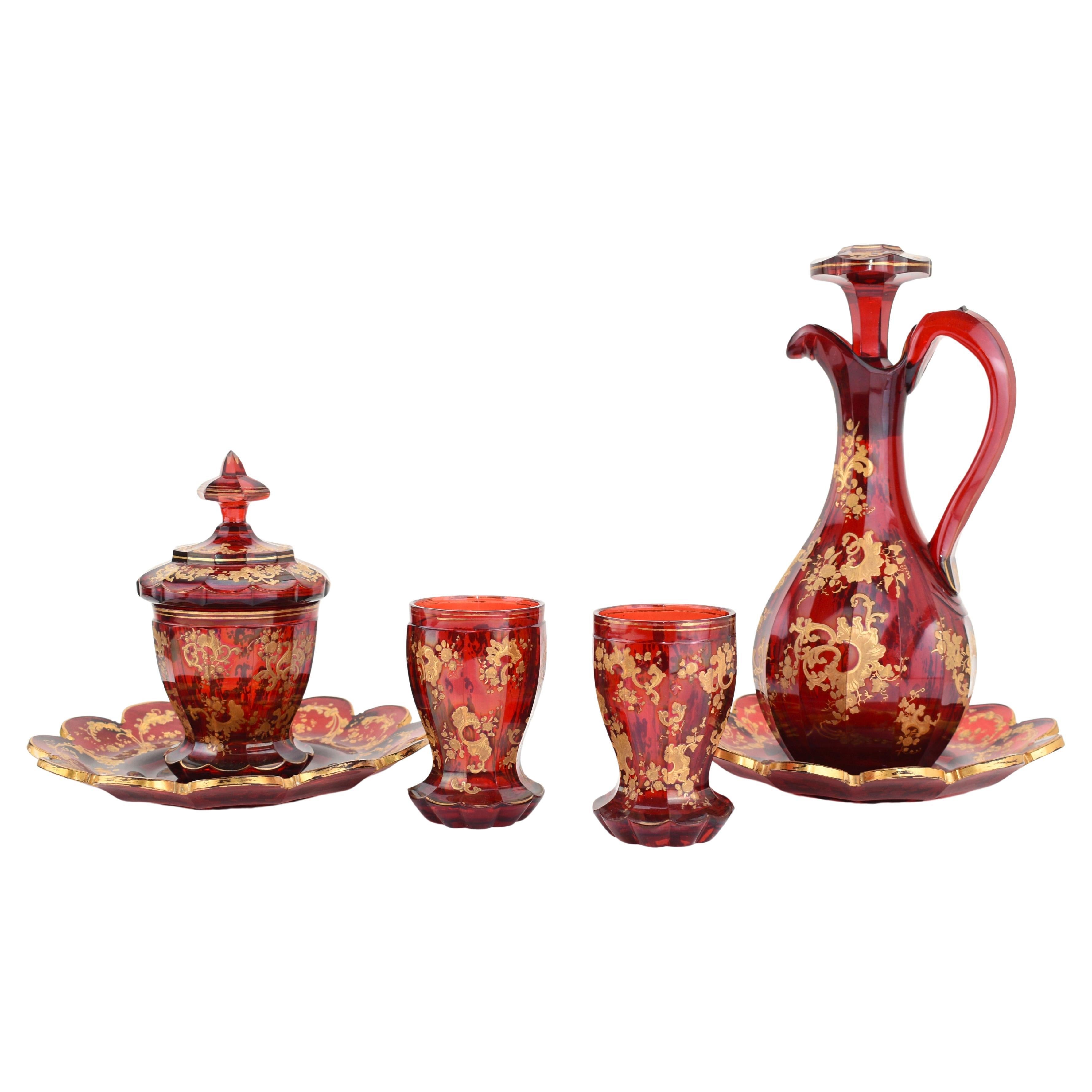 A superb and rare Bohemian gilt and enamelled ruby glass drinks set. This complete set includes a large ewer, two tumblers, a covered sugar bowl, and two circular trays, each meticulously adorned with scrolling foliate decoration.

Made from deep