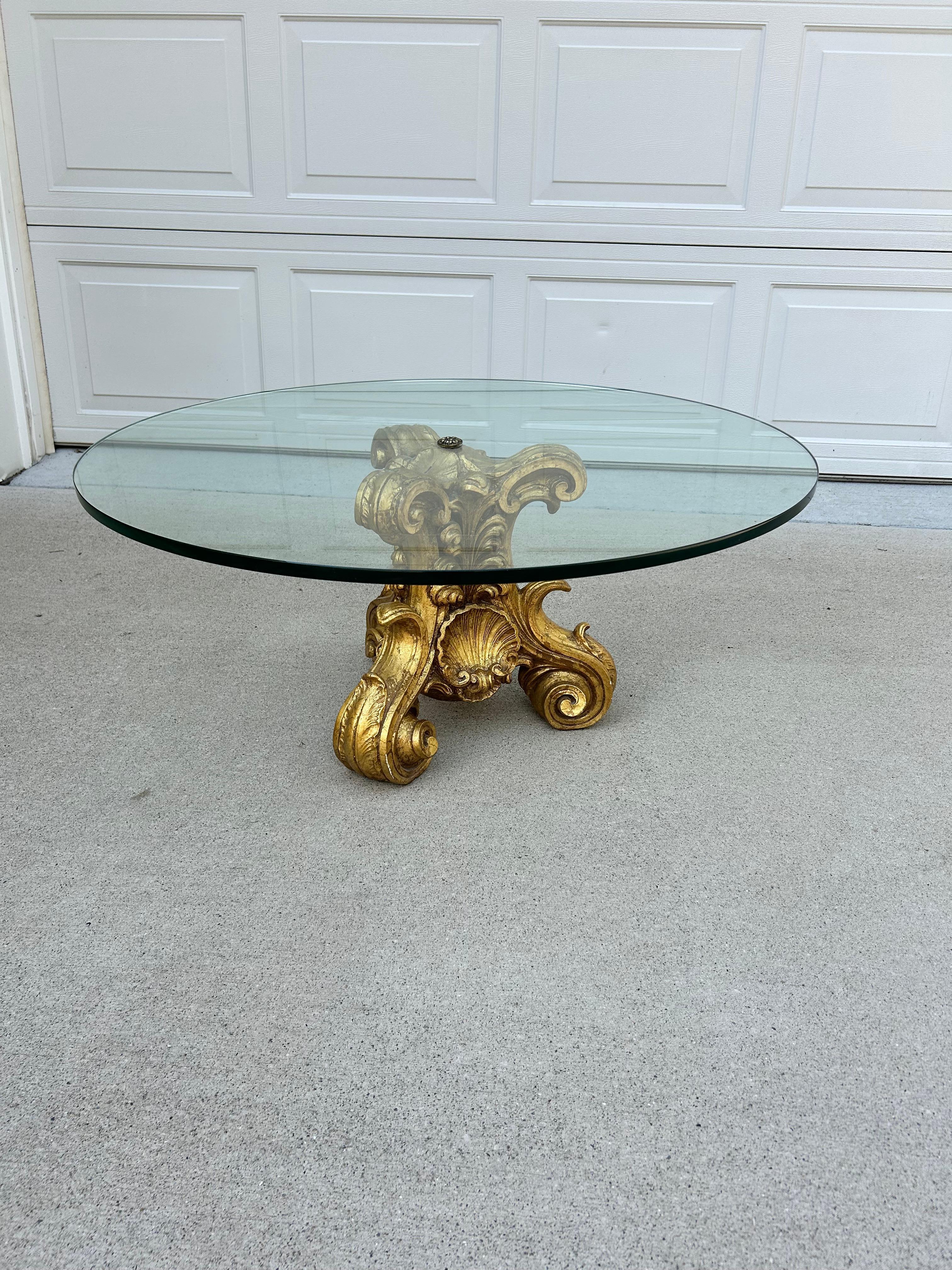 This Early 1900s Hollywood Regency Giltwood, Glass and a touch of Brass Coffee Table exudes opulence and glamour.  Its slightly tinted, glass top offers a sleek surface and is attached perfectly to the giltwood hand carved frame by a decorative