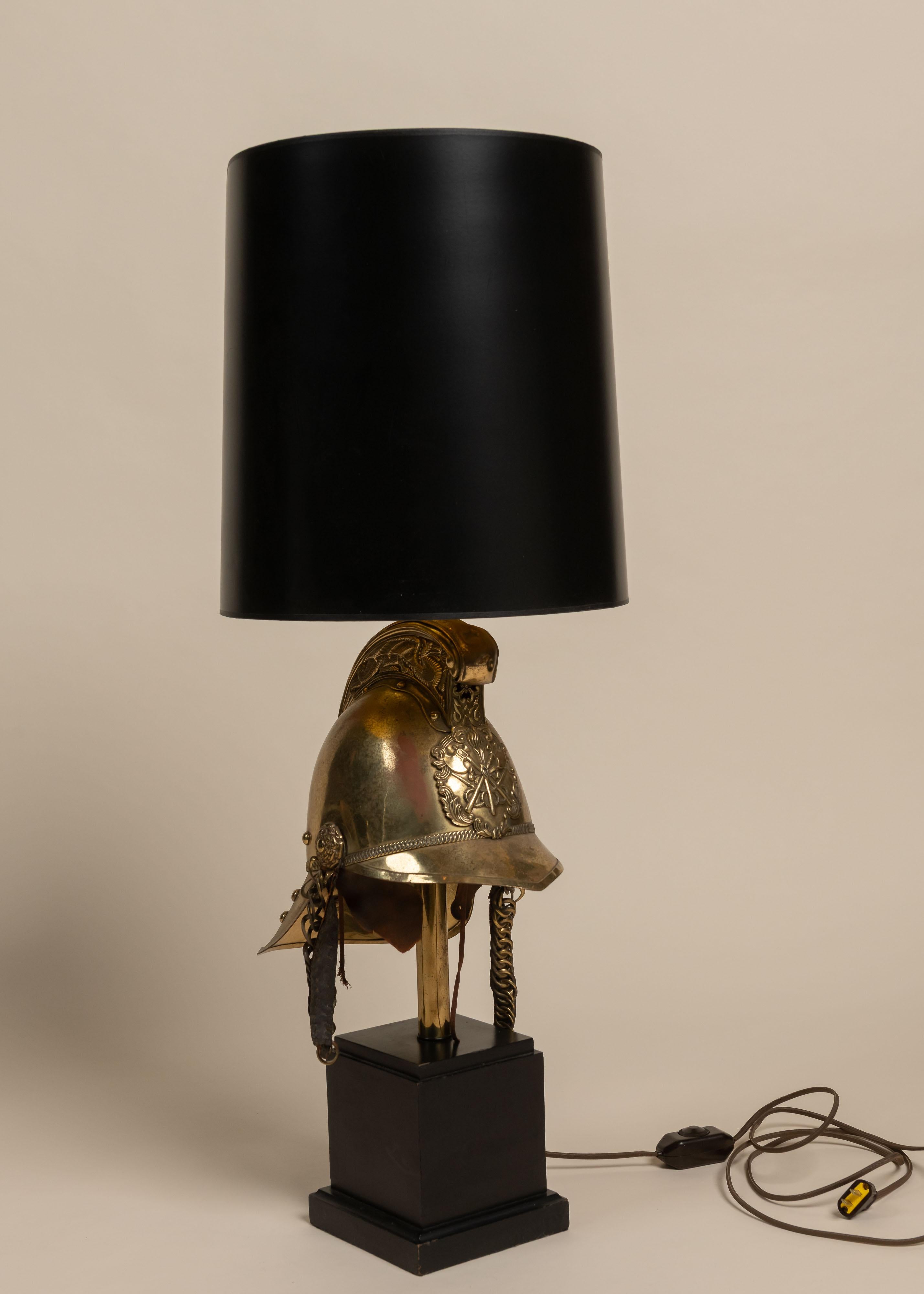Esta espectacular lámpara de sobremesa tiene una base escultural de un casco de la Brigada de Bomberos Británica, con detalles en relieve y borlas laterales. Se asienta sobre un pedestal de madera negra y está coronada por una alta pantalla